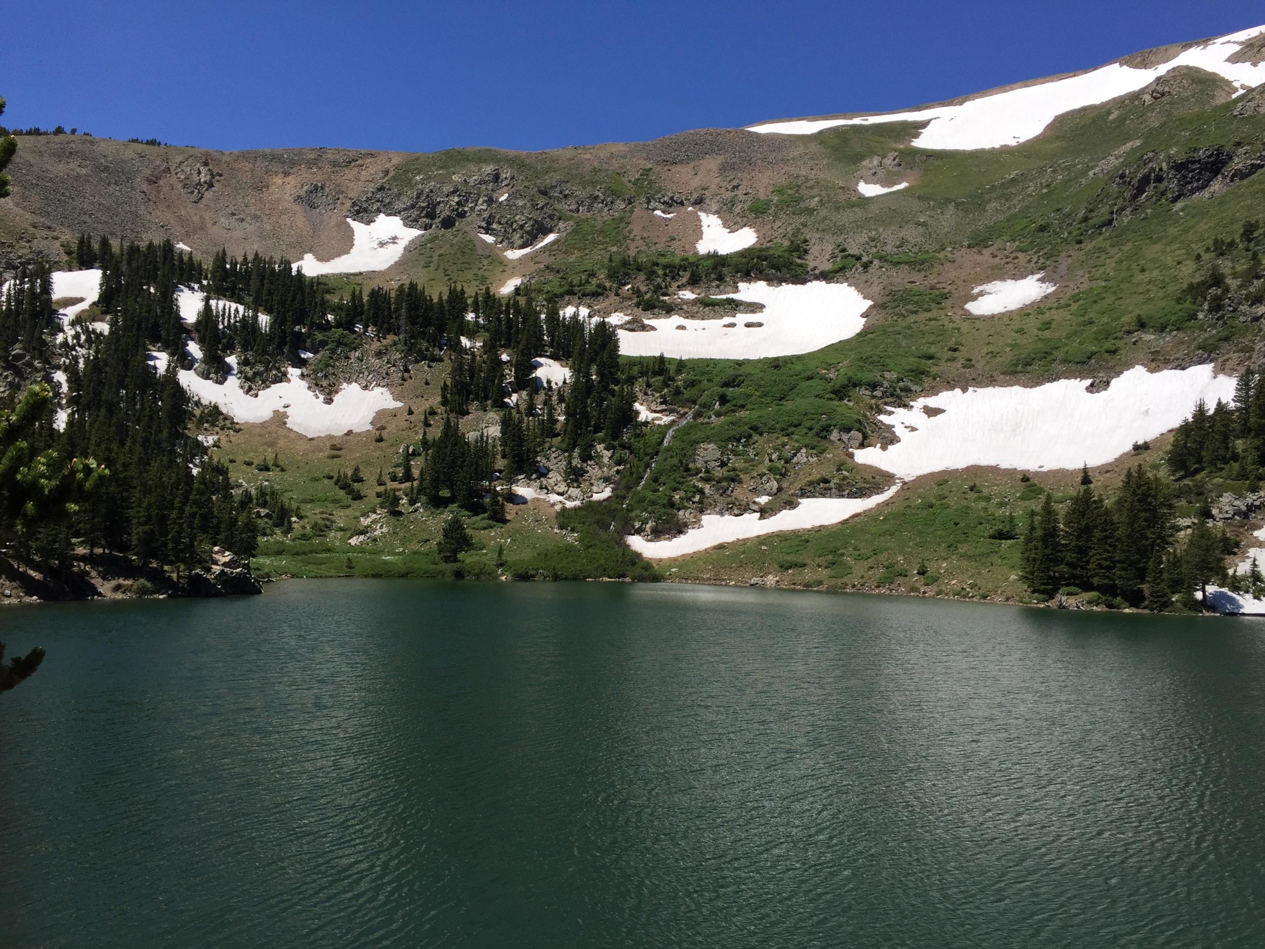 A tranquil alpine lake surrounded by lush greenery and rocky hills, featuring patches of snow on the mountainside. The sky is bright blue, reflecting on the calm water of the lake. Lost Lake to East Fork Loop mountain bike trail.