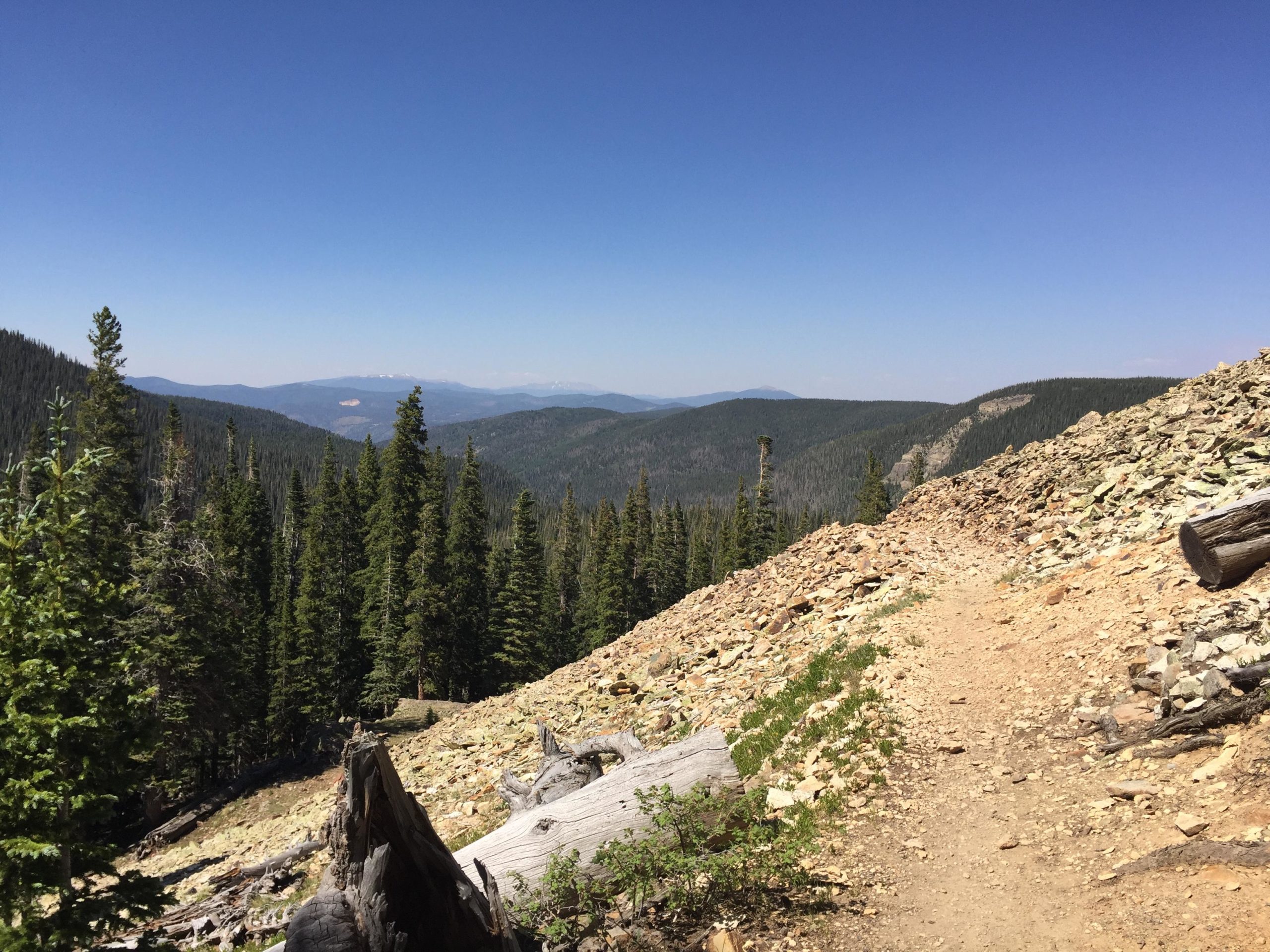 A scenic view of a mountainous landscape featuring rolling hills covered with lush green trees under a clear blue sky. A rocky path winds through the foreground, leading into the distance where more mountains are visible. Lost Lake to East Fork Loop mountain bike trail.