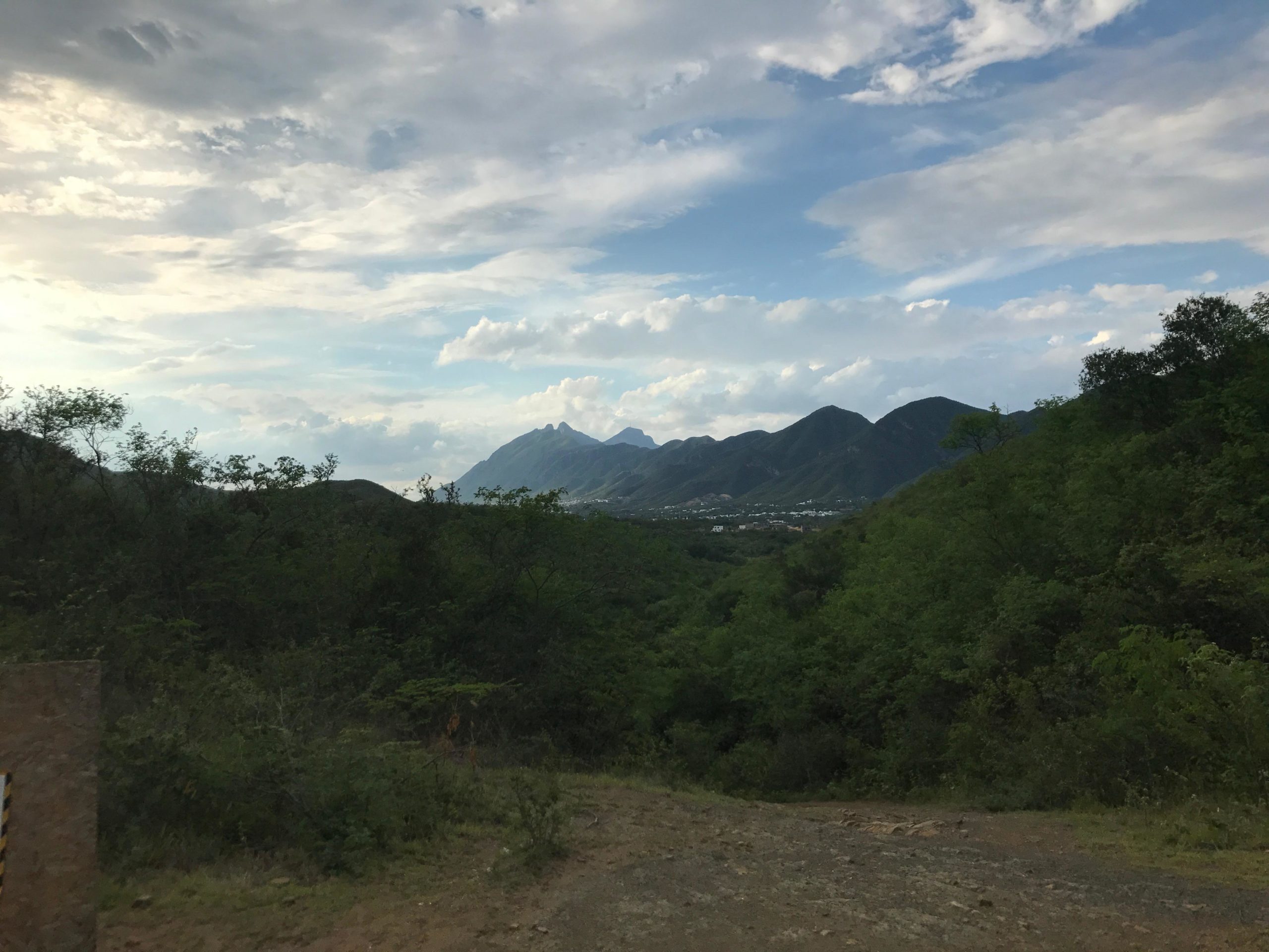 A scenic view of lush green hills leading to distant mountains under a partly cloudy sky. The foreground features a dirt path and patches of vegetation, while the mountains in the background rise dramatically against the sky, creating a serene landscape. la ilusion mountain bike trail.