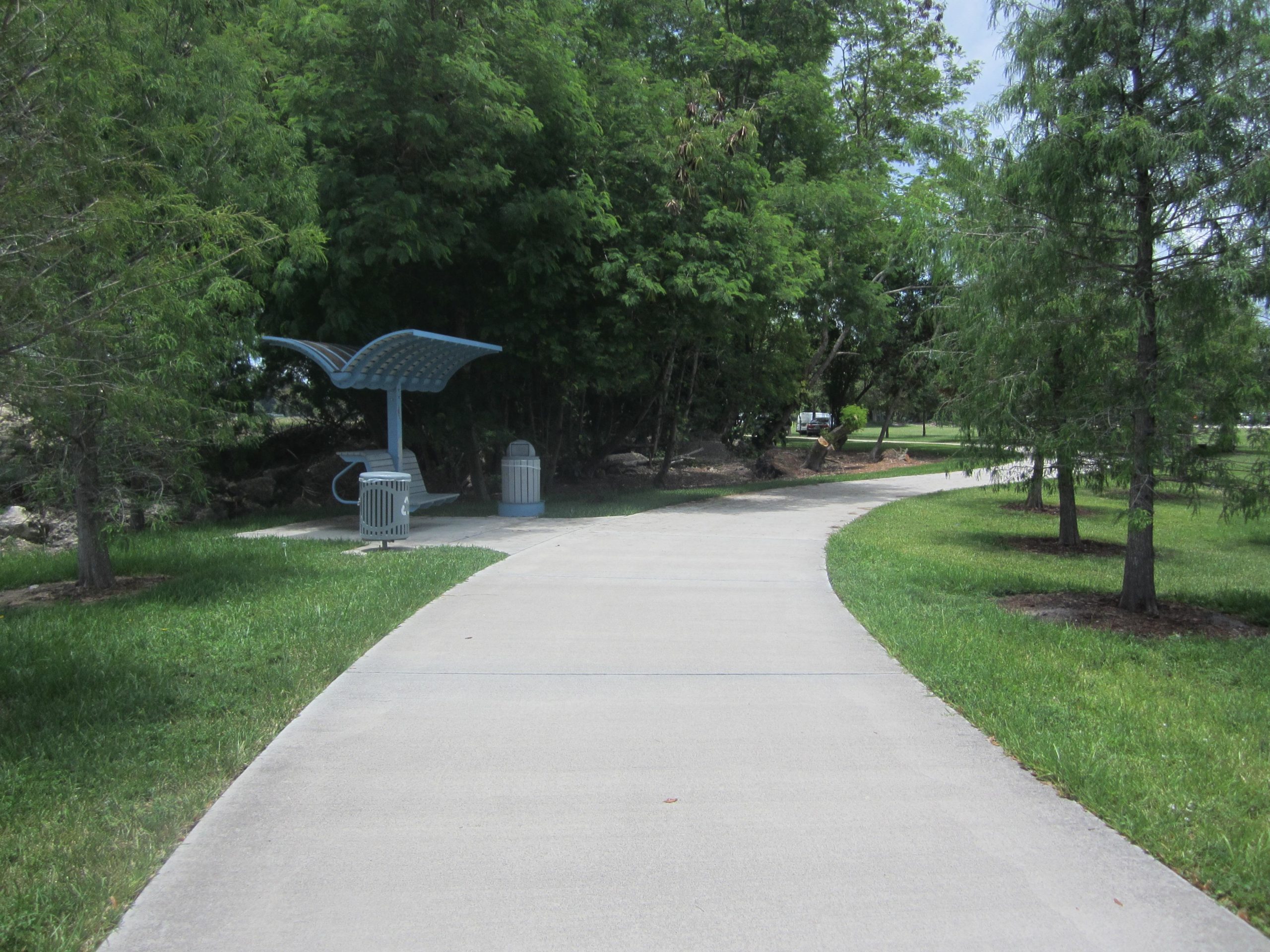 A curving pathway through a green, lush park, lined with trees on both sides. A modern blue bench with a canopy is positioned beside a trash bin to the left of the path. The scene is bright and inviting, suggesting a peaceful outdoor space for walking or resting. New River Greenway mountain bike trail.