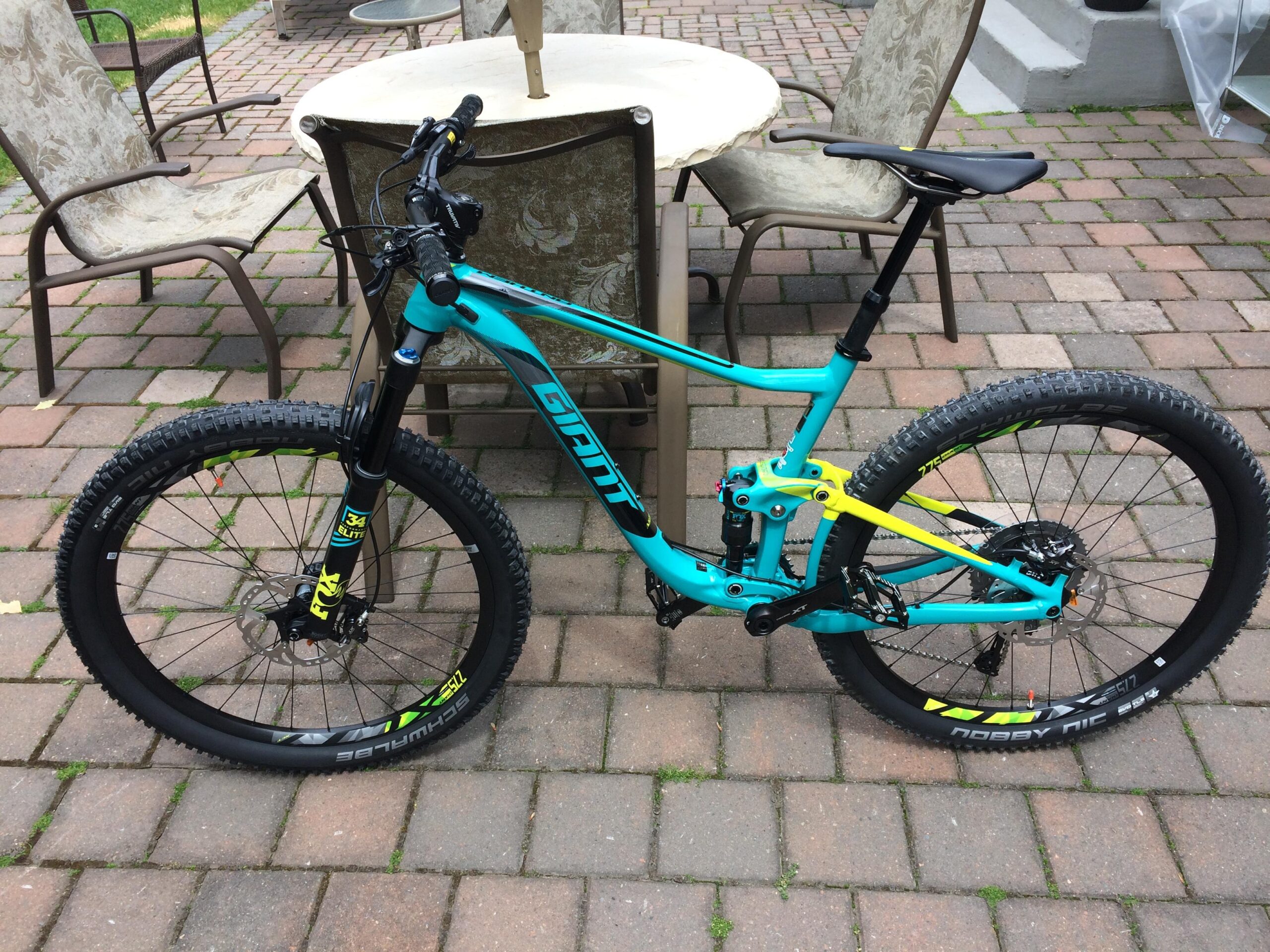 Giant Anthem SX 27.5: A bright turquoise mountain bike with black and yellow accents parked on a patterned brick patio, surrounded by outdoor furniture. The bike features a full suspension, thick tires, and a sleek frame design.