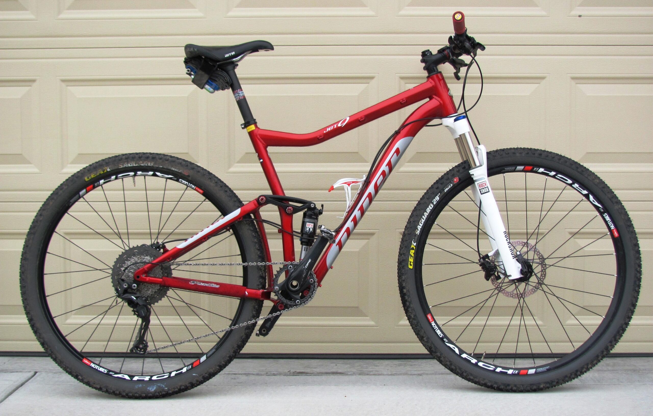 Niner Jet 9: A red and white mountain bike with dual suspension, featuring wide tires and disc brakes, parked in front of a beige garage door. The bike has a sleek design, with a prominent frame, and is equipped with quality components suitable for off-road riding.