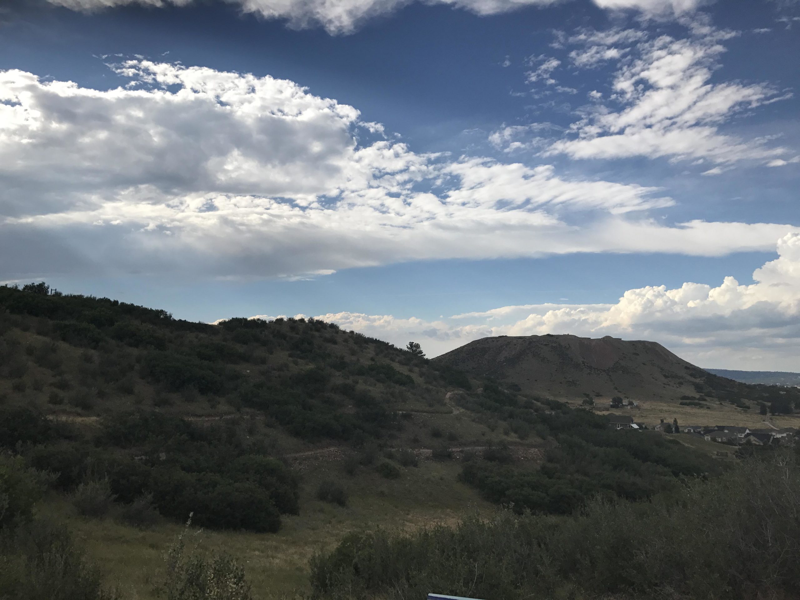 A scenic view of rolling hills under a partly cloudy sky, with green vegetation covering the slopes. In the distance, a prominent hilltop rises against a backdrop of blue sky and white clouds, creating a tranquil natural landscape. Phillip S. Miller Park mountain bike trail.