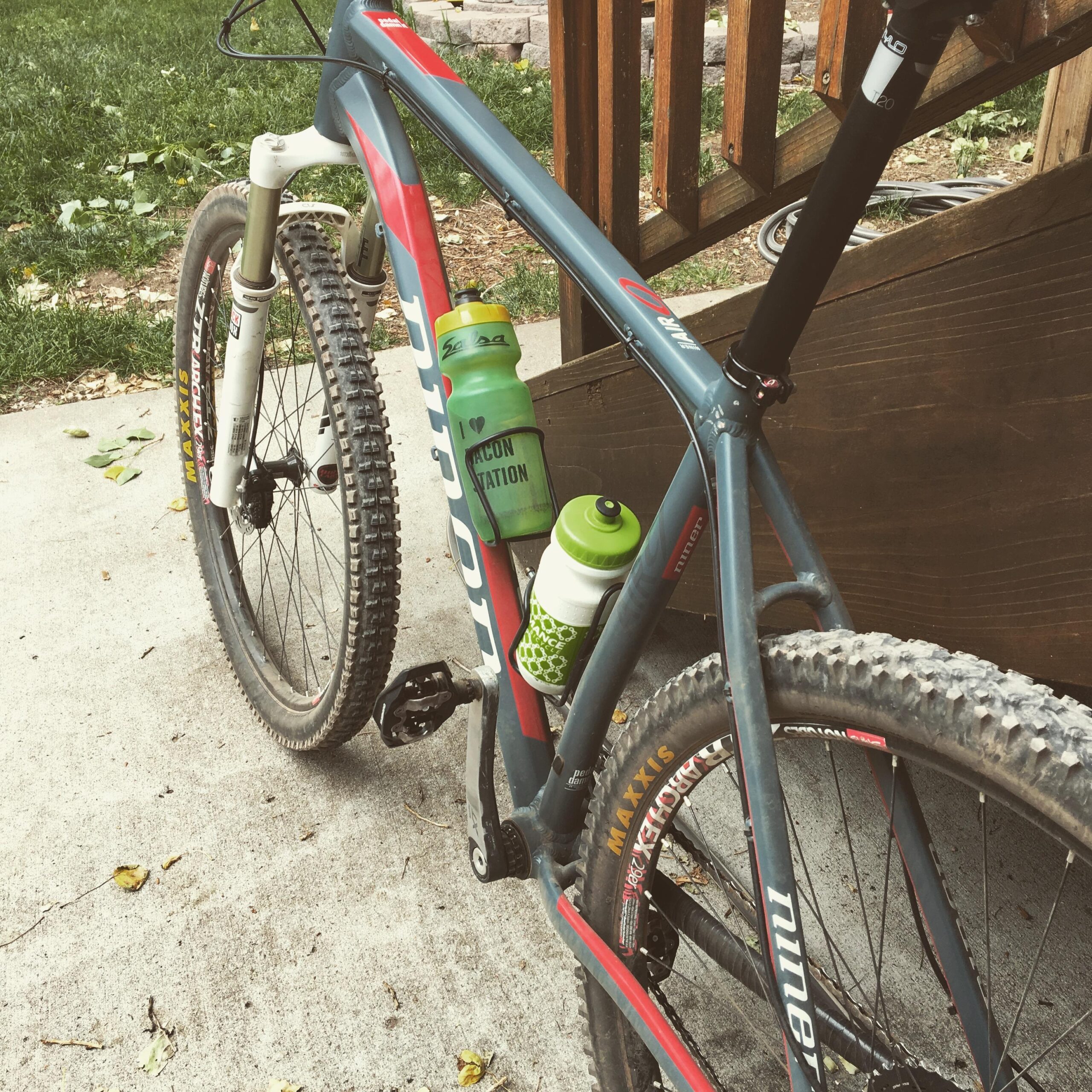 Niner Air 9: A close-up view of a mountain bike parked on a concrete surface, showcasing its frame, tires, and two water bottles attached to the bike. The bike features a gray frame with red accents, rugged tires, and a visible pedal. The water bottles are green and white, with one labeled "I love Falcon Nation." In the background, there are patches of grass and a wooden structure.