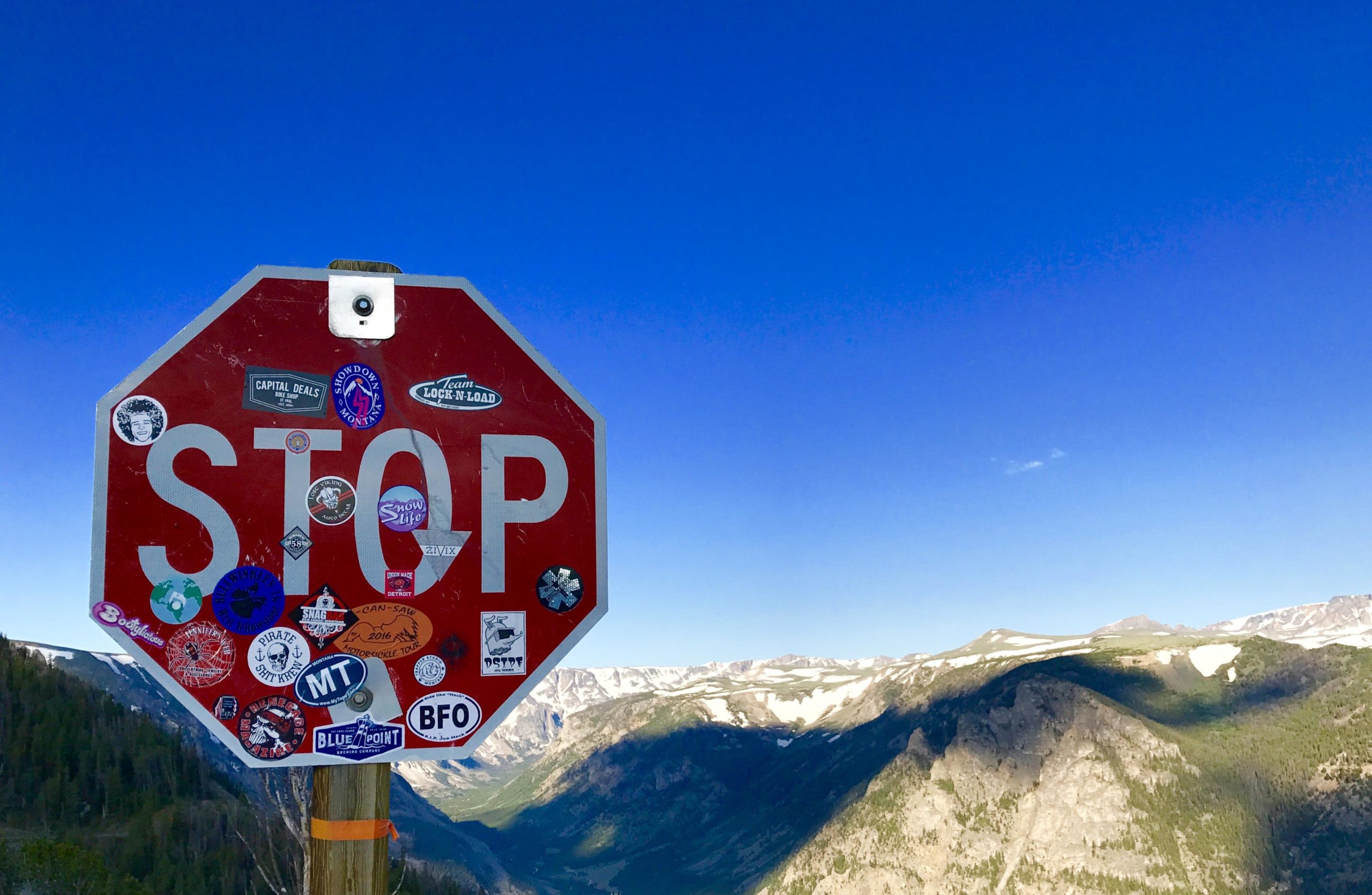 A weathered stop sign covered in various stickers, set against a clear blue sky and mountainous landscape. The foreground features the stop sign prominently, while the background displays lush green valleys and snow-capped mountains. Line Creek Plateau mountain bike trail.