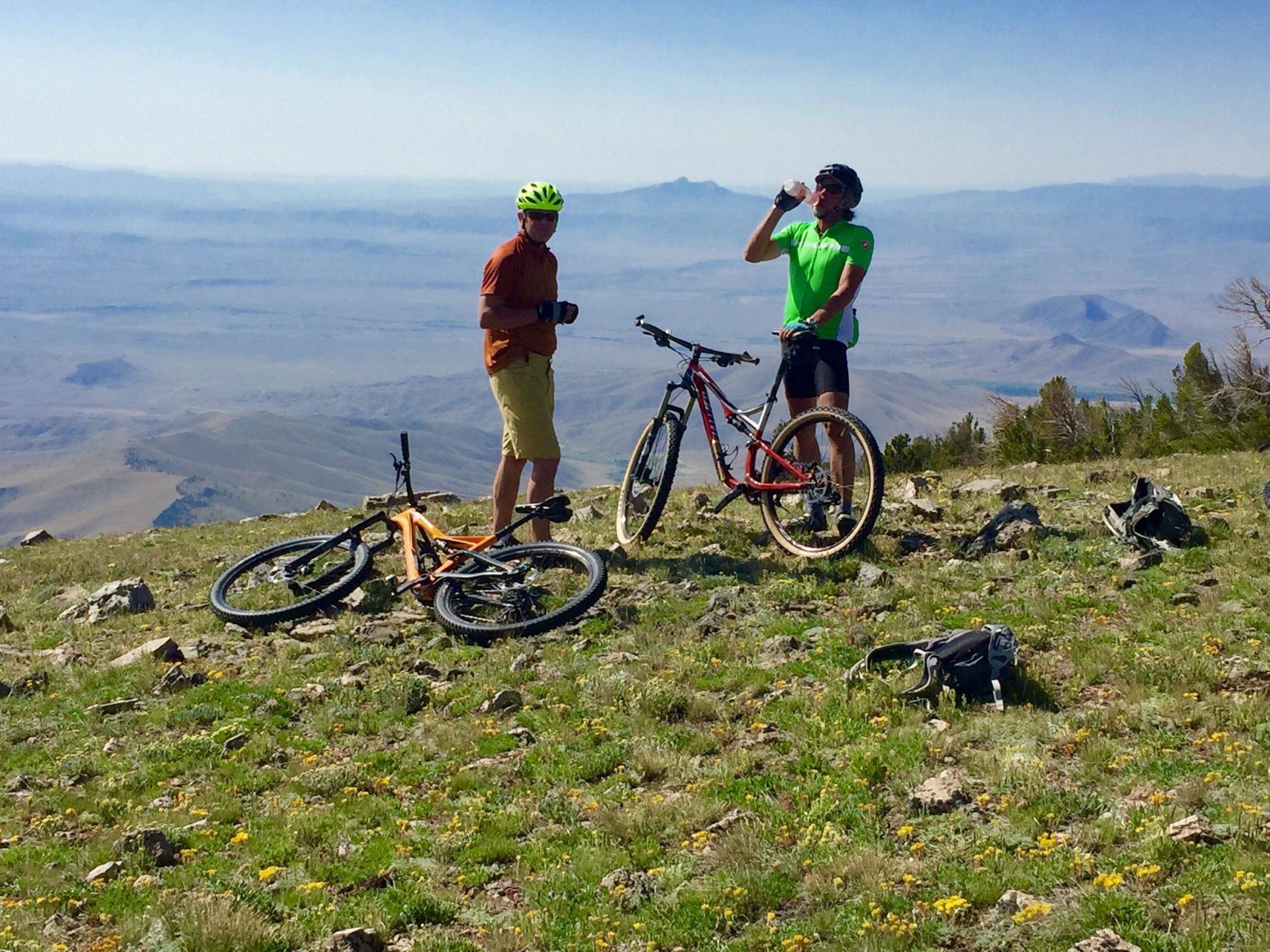 Two mountain bikers resting on a grassy, rocky landscape at a high elevation. One biker, wearing a bright green jersey and drinking water, stands next to his bike, while the other, in an orange shirt and shorts, is holding a camera. In the background, expansive mountains and valleys create a scenic view under a clear blue sky. Line Creek Plateau mountain bike trail.