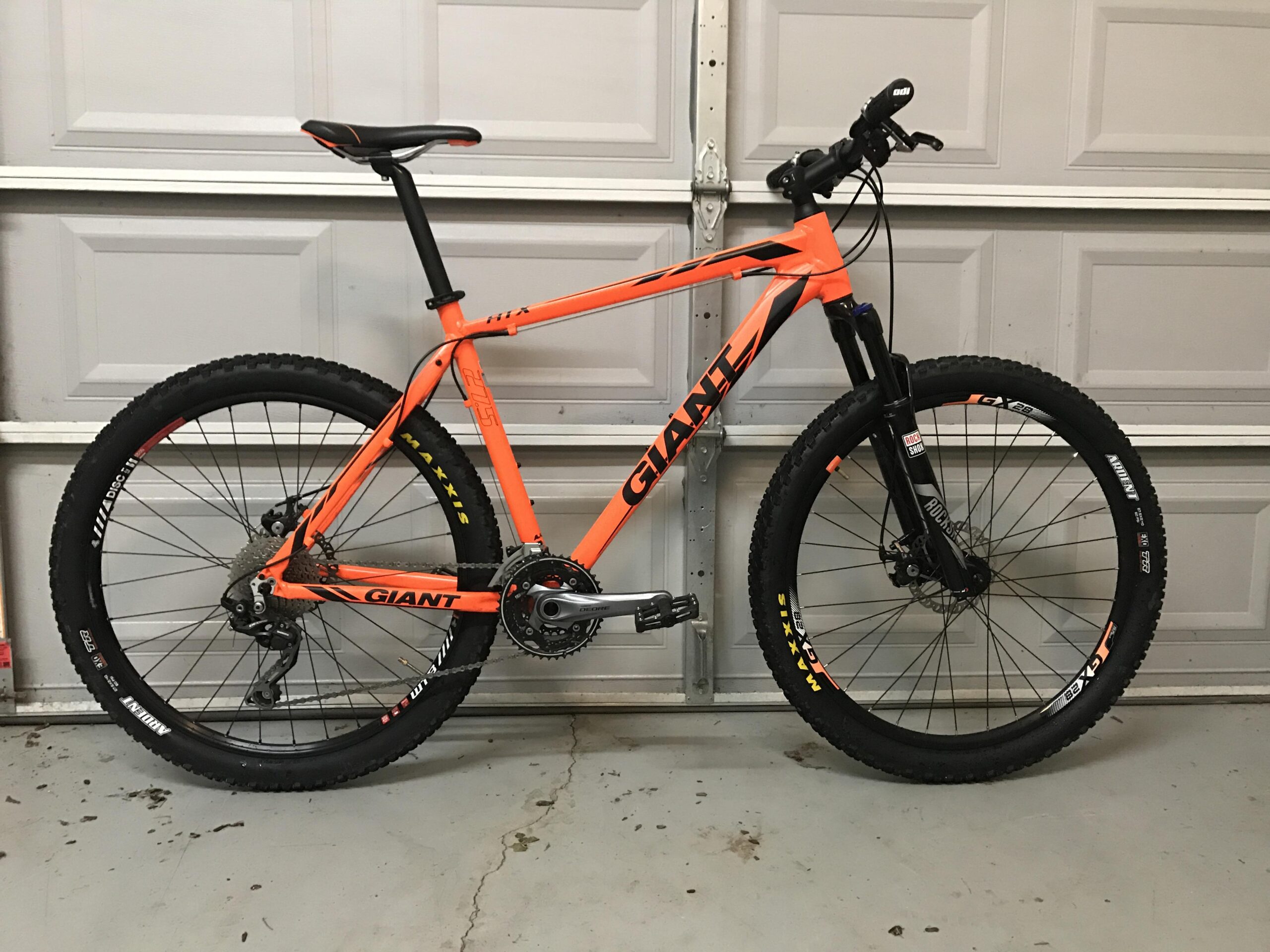 Giant ATX 27.5 2: Alt text: A bright orange mountain bike with black details and rims, featuring knobby tires and a modern frame design, positioned against a garage backdrop.