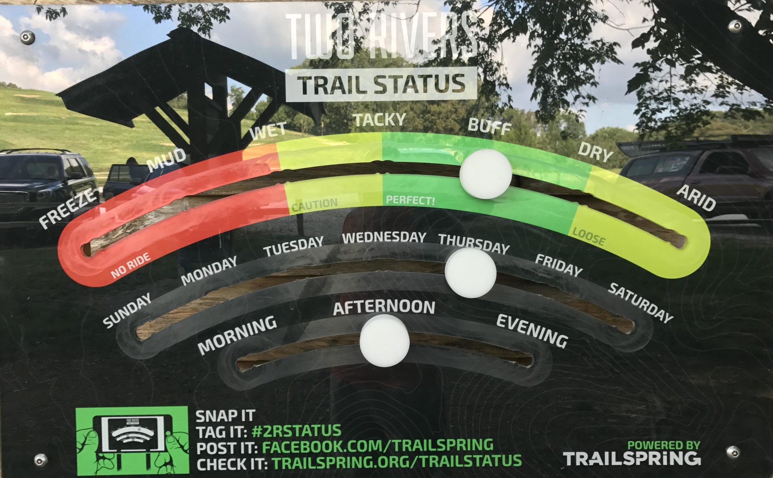 A trail status board for Two Rivers, displaying conditions for various days of the week. The board features color-coded indicators ranging from "Freeze" (red, no ride) to "Arid" (green, loose). Markers note trail conditions for each day and time of day, including "Wet," "Tacky," "Buff," "Dry," and "Perfect." The bottom section encourages users to engage through social media, with instructions for tagging and posting updates. Two Rivers Bike Park mountain bike trail.
