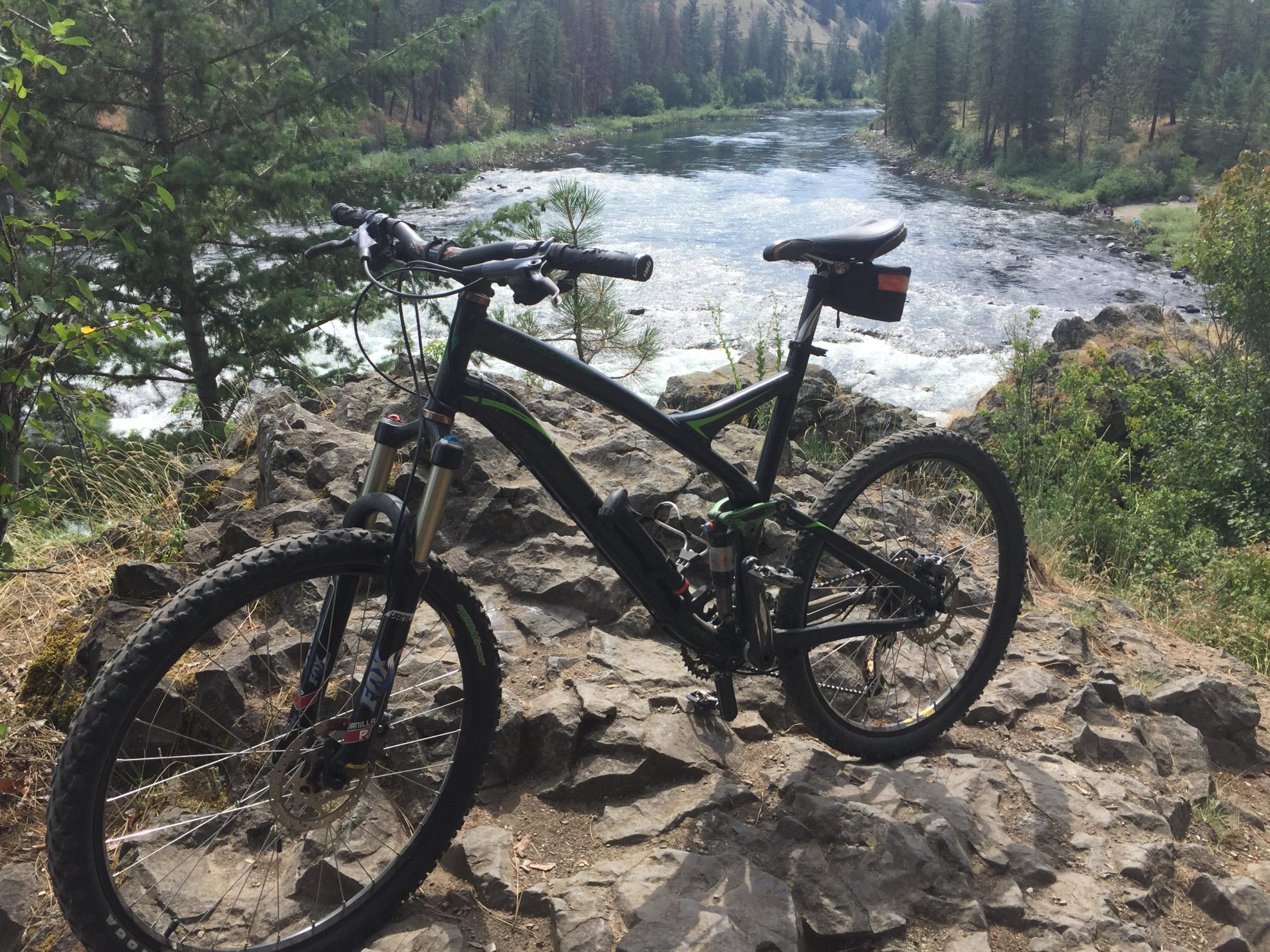 A black mountain bike resting on rocky terrain with a river flowing in the background, surrounded by lush trees and greenery. The scene captures a peaceful outdoor setting, ideal for biking enthusiasts. Riverside State Park mountain bike trail.