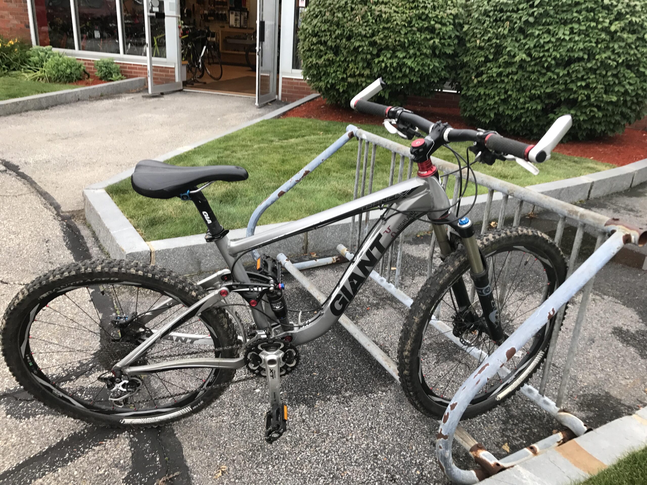 Giant Trance X1: A gray mountain bike is secured to a bike rack outside a shop. The bike features thick tires, a sturdy frame, and black grips on the handlebars. In the background, there is a shop entrance with glass doors, and greenery surrounds the area.