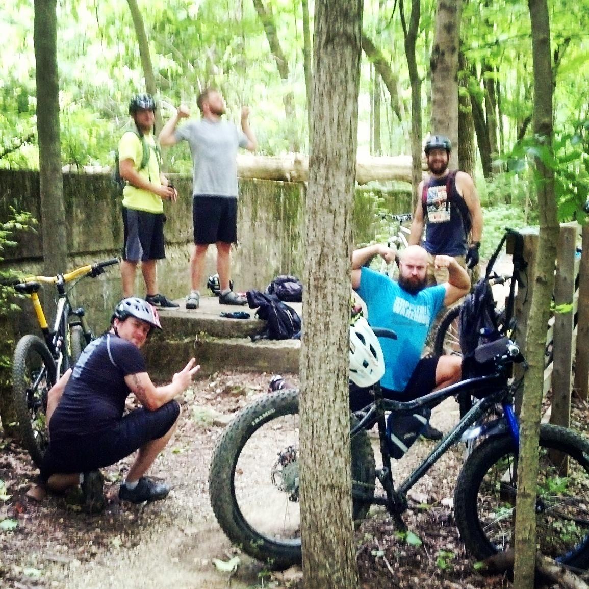 A group of five cyclists taking a break in a lush, green forest setting. Two men stand on a stone platform, with one raising his arms in celebration and the other smiling at the camera. In the foreground, a man in a black shirt poses with a peace sign while kneeling beside his mountain bike. Another bike is parked nearby, partially obscured by a tree. The scene conveys a sense of camaraderie and enjoyment of outdoor activities. Fort Harrison State Park mountain bike trail.