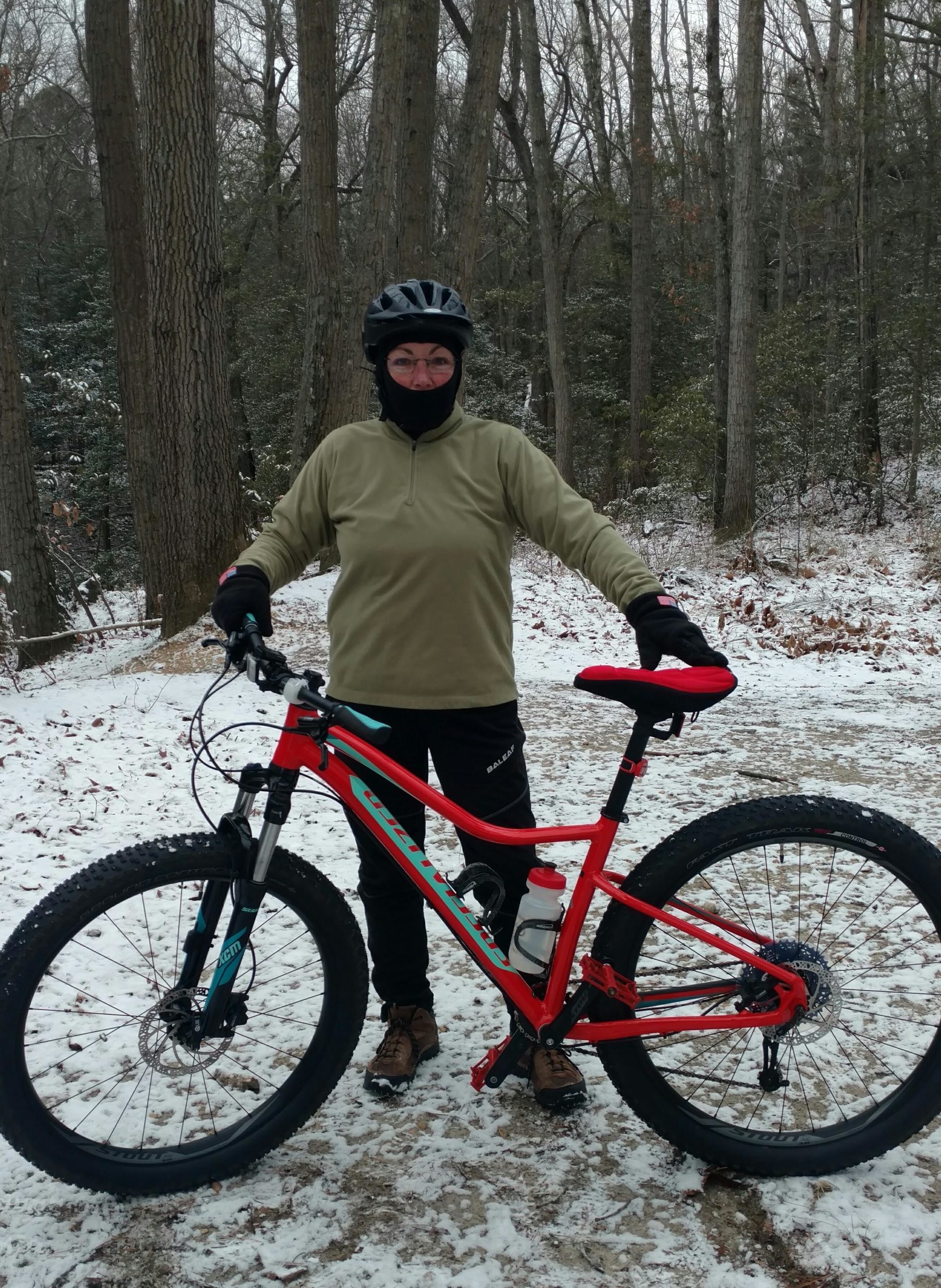 A person stands beside a red mountain bike on a snowy path surrounded by trees. They are wearing a helmet, a thermal neck gaiter, and gloves, indicating cold weather. The ground is covered with a light layer of snow, and the atmosphere is serene, showcasing a wintery outdoor biking environment. Camden County College mountain bike trail.