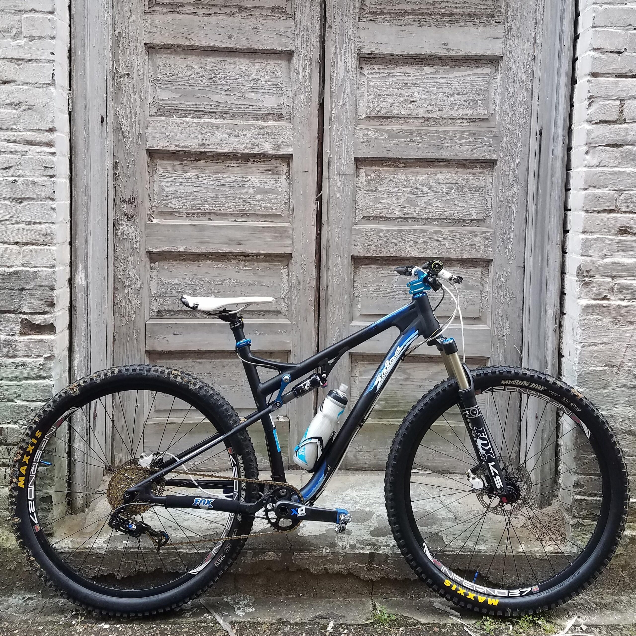 Salsa Spearfish: A mountain bike with a blue and black frame is positioned in front of an old wooden door. The bike features a water bottle attached to the frame and has rugged tires designed for off-road riding. The background consists of weathered brick and an aged door with multiple panels.