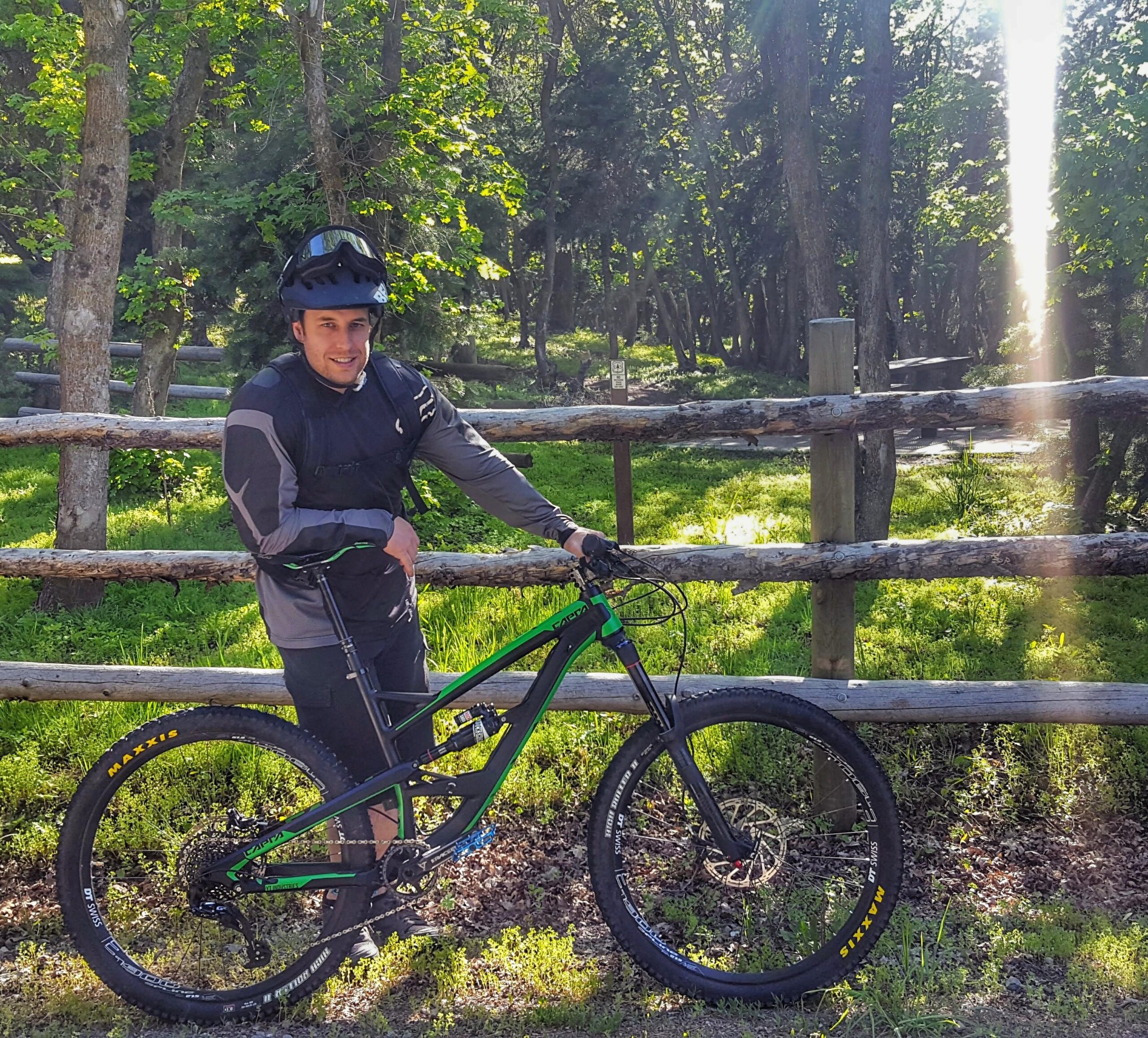 YT Capra: A person wearing a helmet and biking gear stands beside a mountain bike in a lush green forest. The bike is black and green with thick tires, and the background features trees and a wooden fence. Sunlight filters through the leaves, creating a bright and inviting atmosphere.