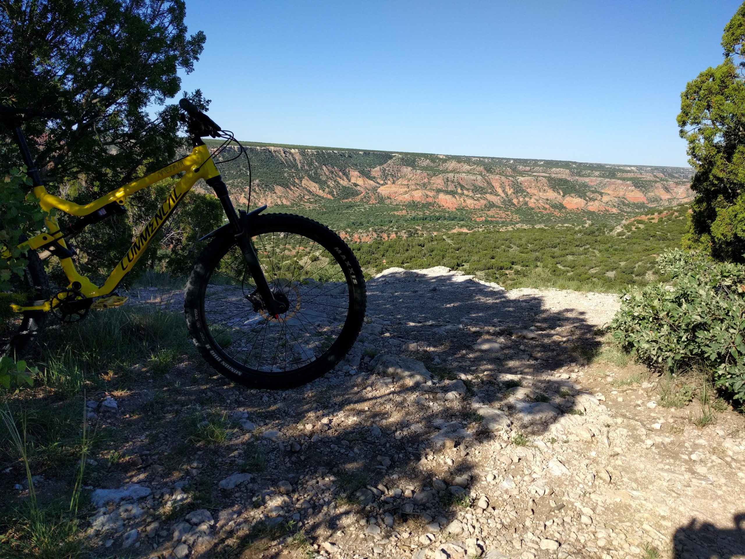 A yellow mountain bike leaning against a tree on a rocky trail, overlooking a scenic view of layered cliffs and greenery under a clear blue sky. Palo Duro Canyon mountain bike trail.