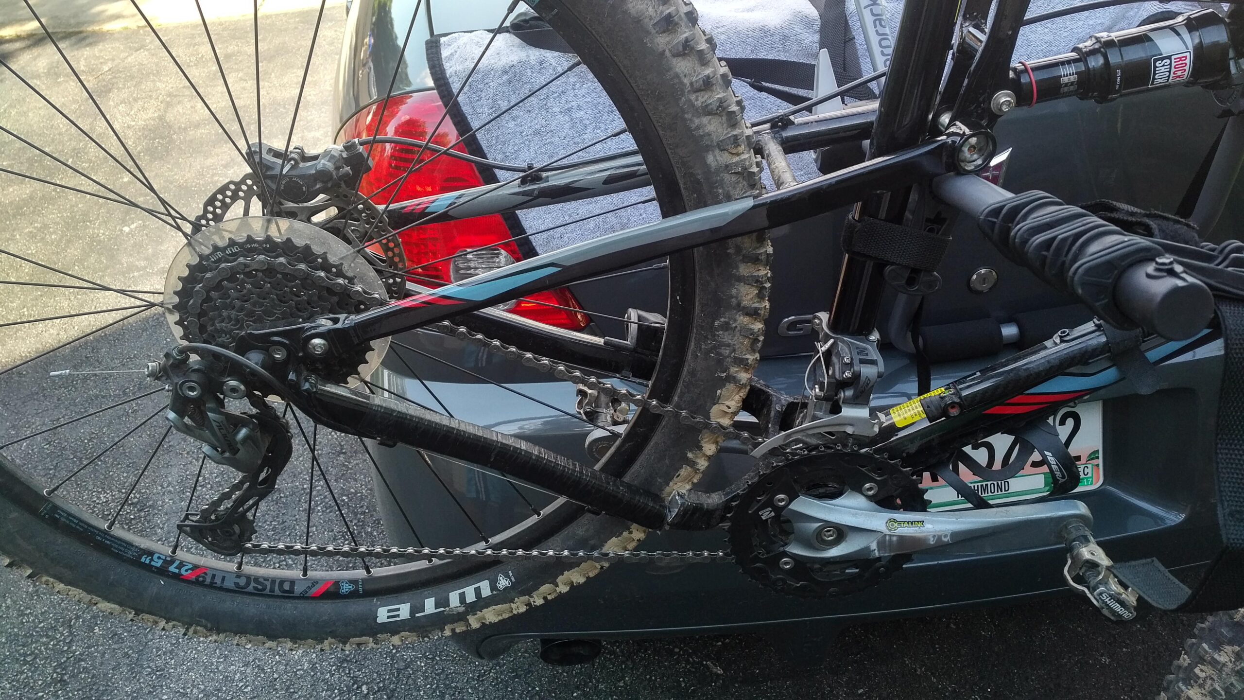 Jamis Dakar XC: A close-up view of the rear section of a mountain bike attached to a vehicle, showcasing the derailleur, rear wheel, and drivetrain components. The bike has visible dirt on the tires, indicating recent use. The background features the rear of a grey car with a partial view of its taillight and a towel on the roof.