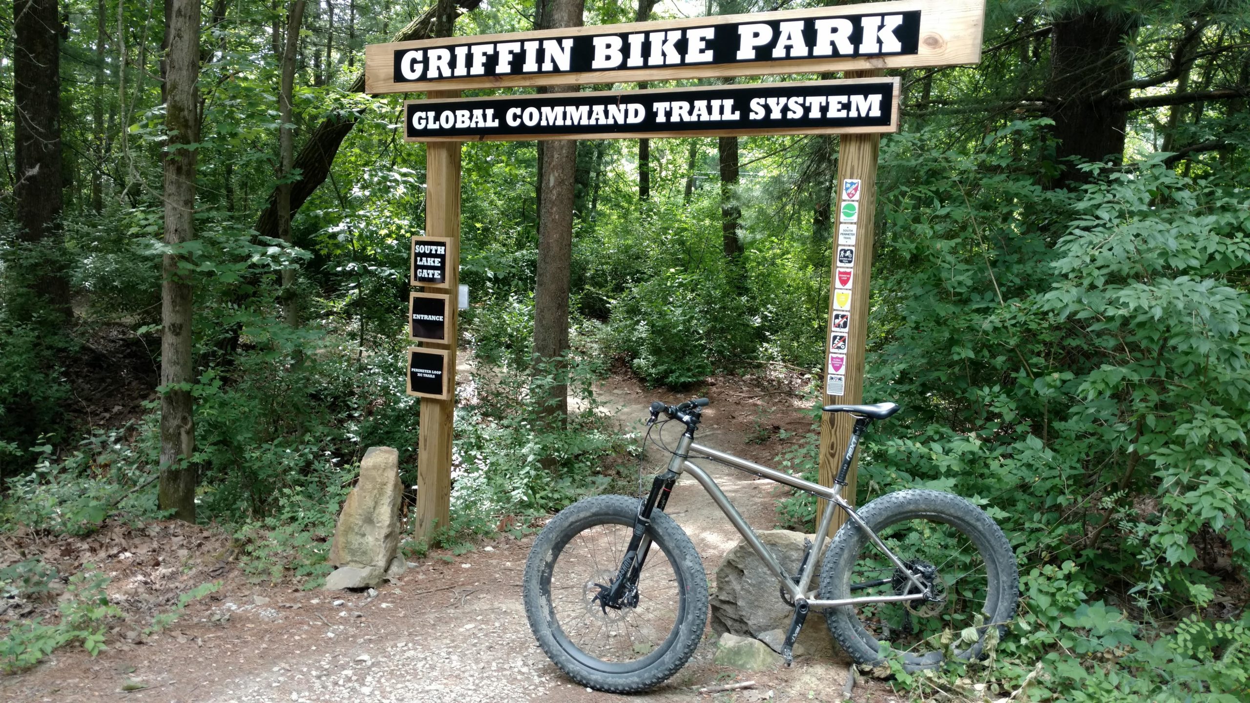 A mountain bike parked next to a wooden sign for Griffin Bike Park, indicating the entrance to the Global Command Trail System. The area is surrounded by dense green trees and foliage, providing a natural setting for outdoor activities. Griffin Bike Park mountain bike trail.