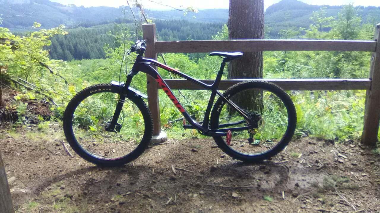 Kona Honzo: A black mountain bike with red accents is parked beside a wooden fence, overlooking a lush landscape of forests and hills under a bright sky. The scene conveys a sense of adventure and outdoor activity.