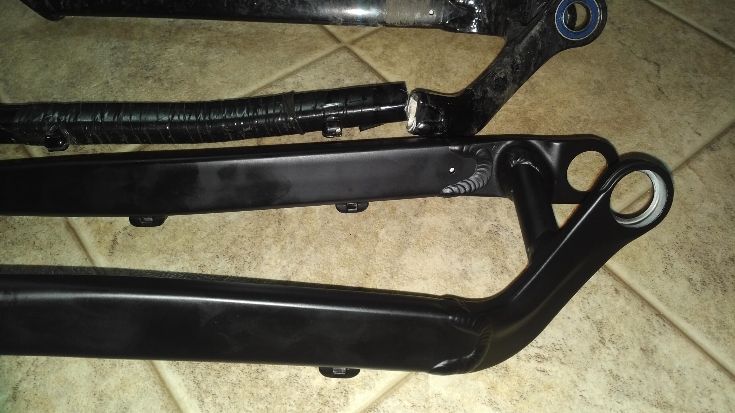 Jamis Dakar XC: Three black suspension links are laid out on a textured, light-colored surface. The top link has a glossy finish, while the middle link is covered with a protective wrapping. The bottom link has a matte finish with visible connection points.