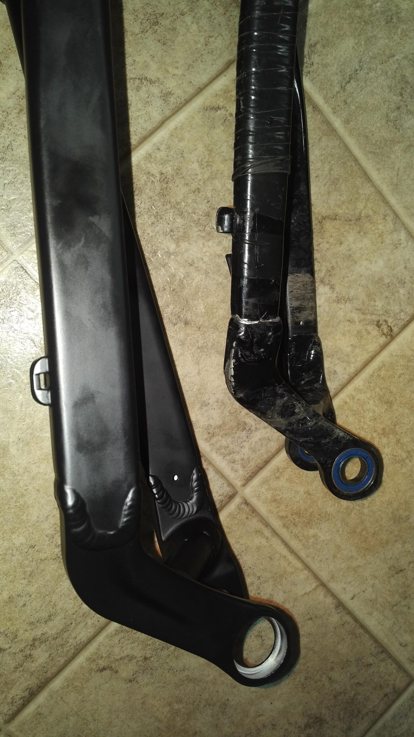 Jamis Dakar XC: Image of two bicycle suspension arms positioned next to each other on a tiled surface. The left arm is sleek and coated in a glossy black finish, while the right arm appears to have signs of wear and is partially wrapped in tape.