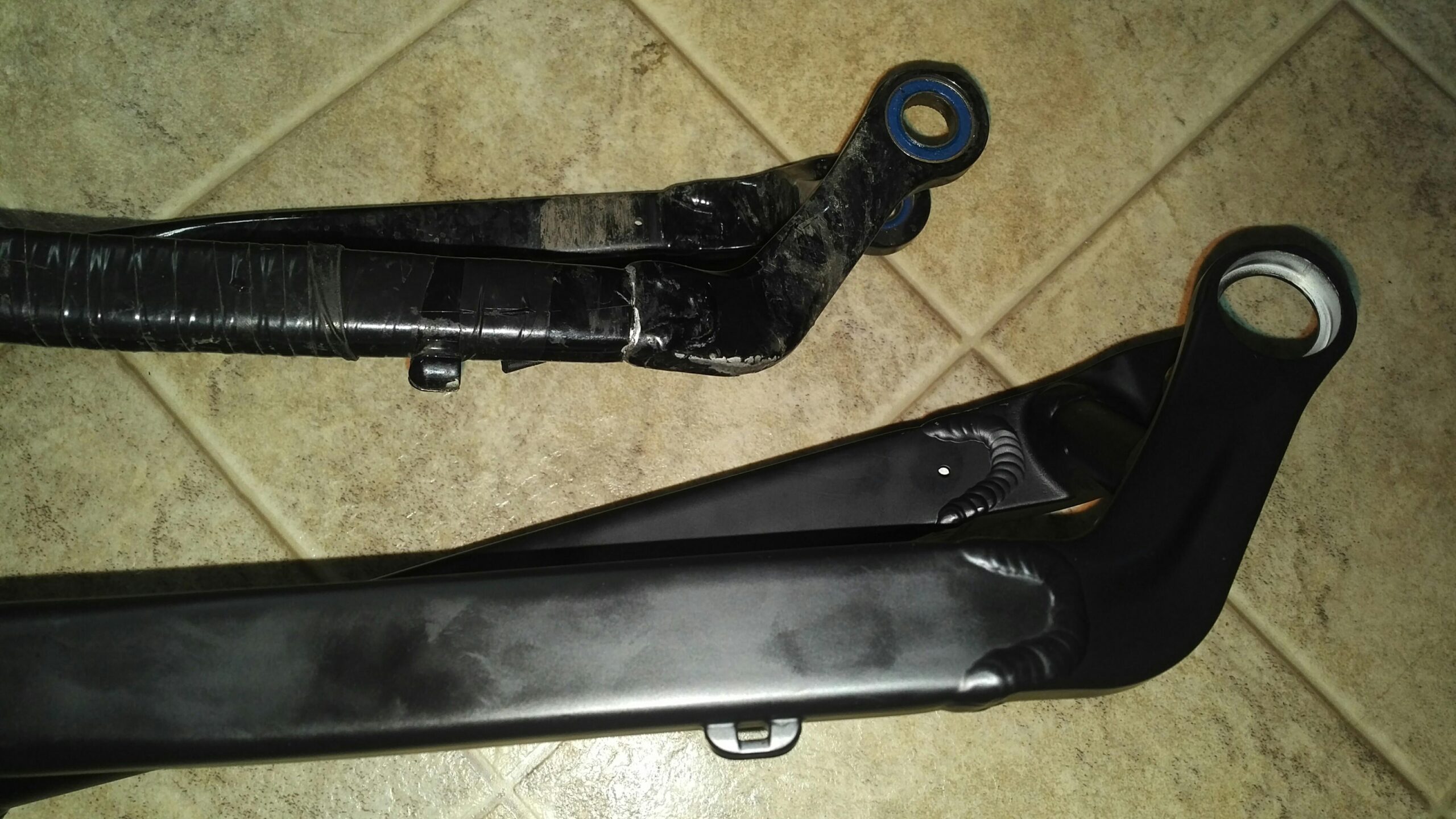 Jamis Dakar XC: Close-up image of two bicycle frame components laid on a tiled surface. The top piece features a rugged, black finish with visible wear, while the bottom piece is sleek and shiny, also in black. Both parts showcase attachment points and structural design elements typical of a bicycle's rear suspension system.