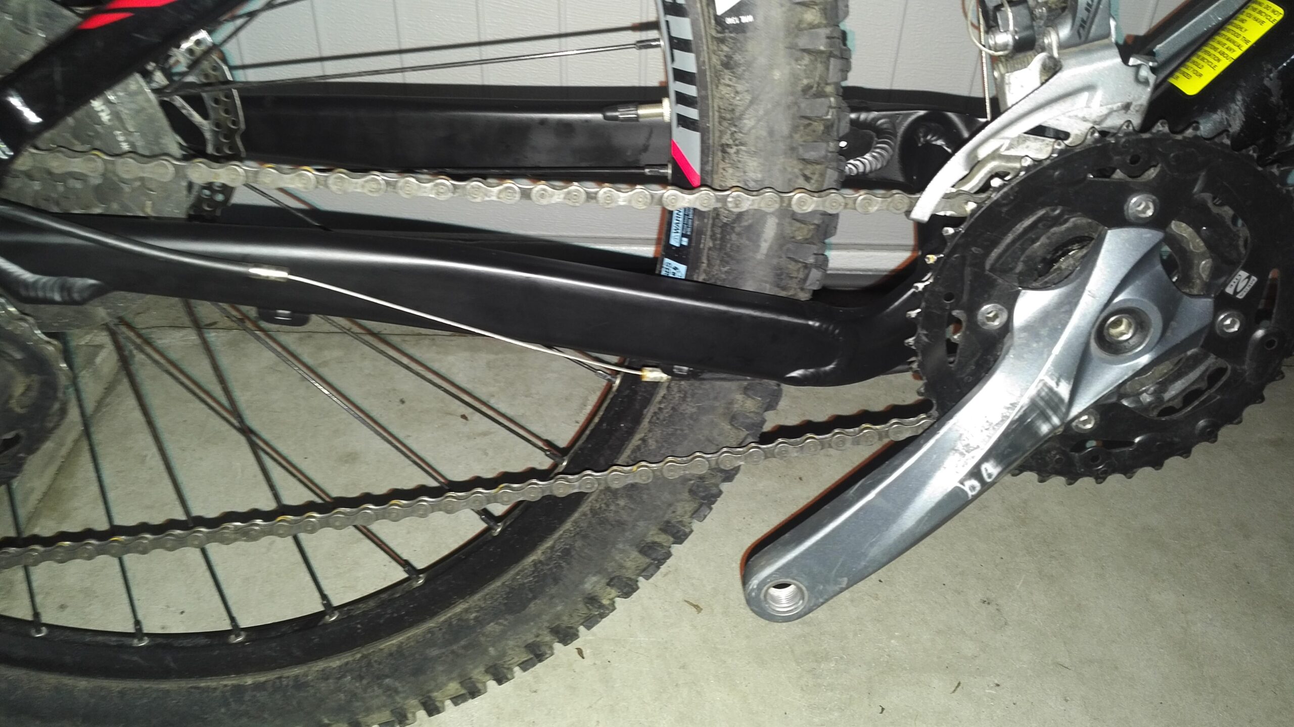 Jamis Dakar XC: Close-up view of a mountain bike's drivetrain, featuring the chain, rear derailleur, and crankset. The bike is positioned on a concrete surface, showcasing the bicycle's components and highlighting the chain's connection to the gear system.