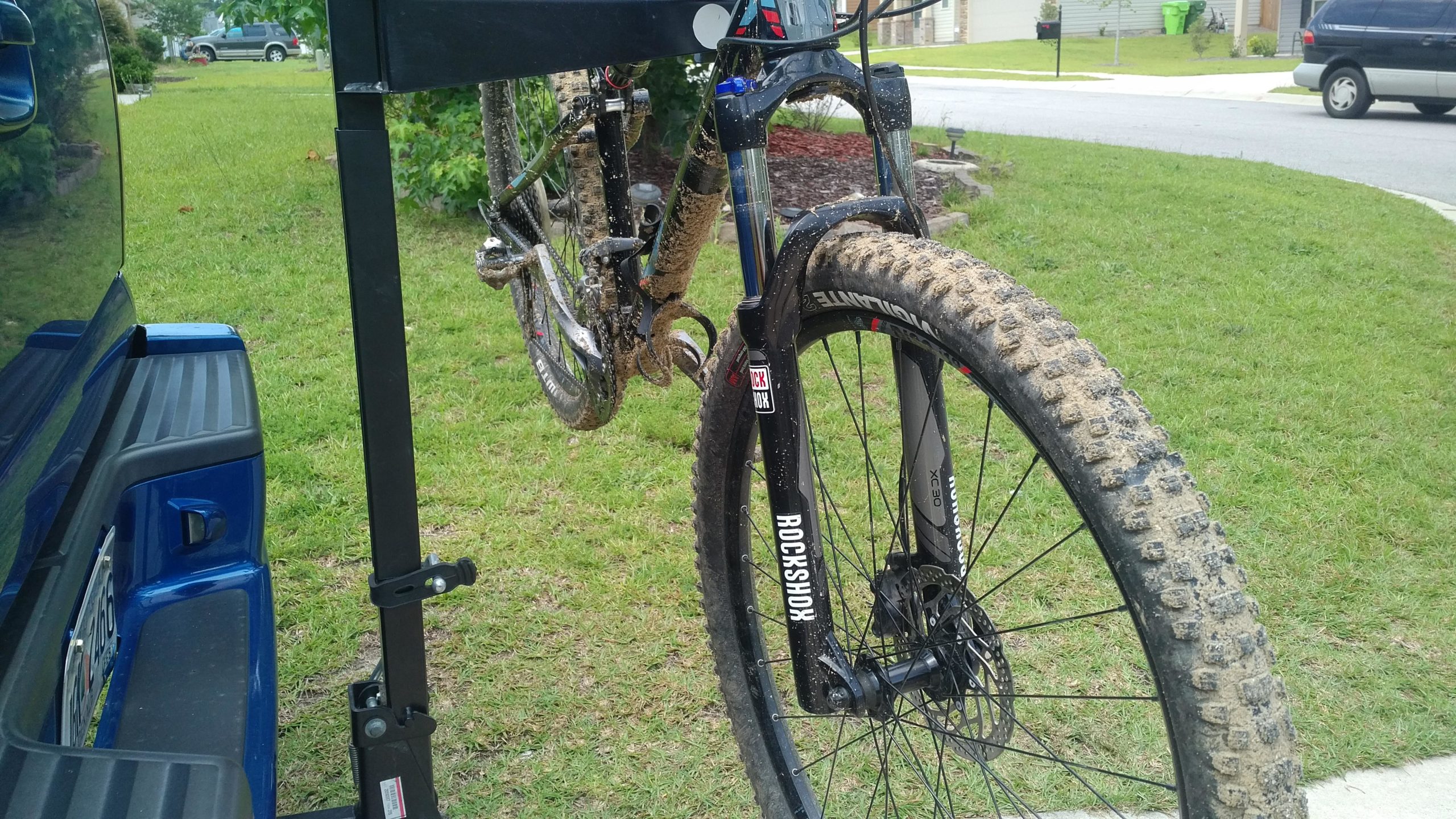 A close-up view of a muddy mountain bike mounted on a hitch rack, beside a blue truck. The bike features thick, textured tires and a visible suspension fork, indicating it's been used on off-road trails. The background includes a grassy area and a suburban street. Horry County Bike Run Park mountain bike trail.