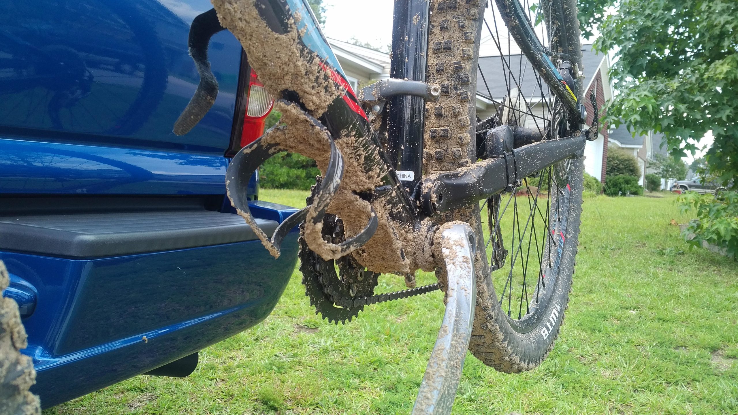 A close-up view of a muddy bicycle positioned next to a blue vehicle. The bike's gears and drivetrain are covered in dirt and mud, indicating it has been recently used in off-road conditions. In the background, a green lawn and part of a house are visible, suggesting a residential setting. Horry County Bike Run Park mountain bike trail.