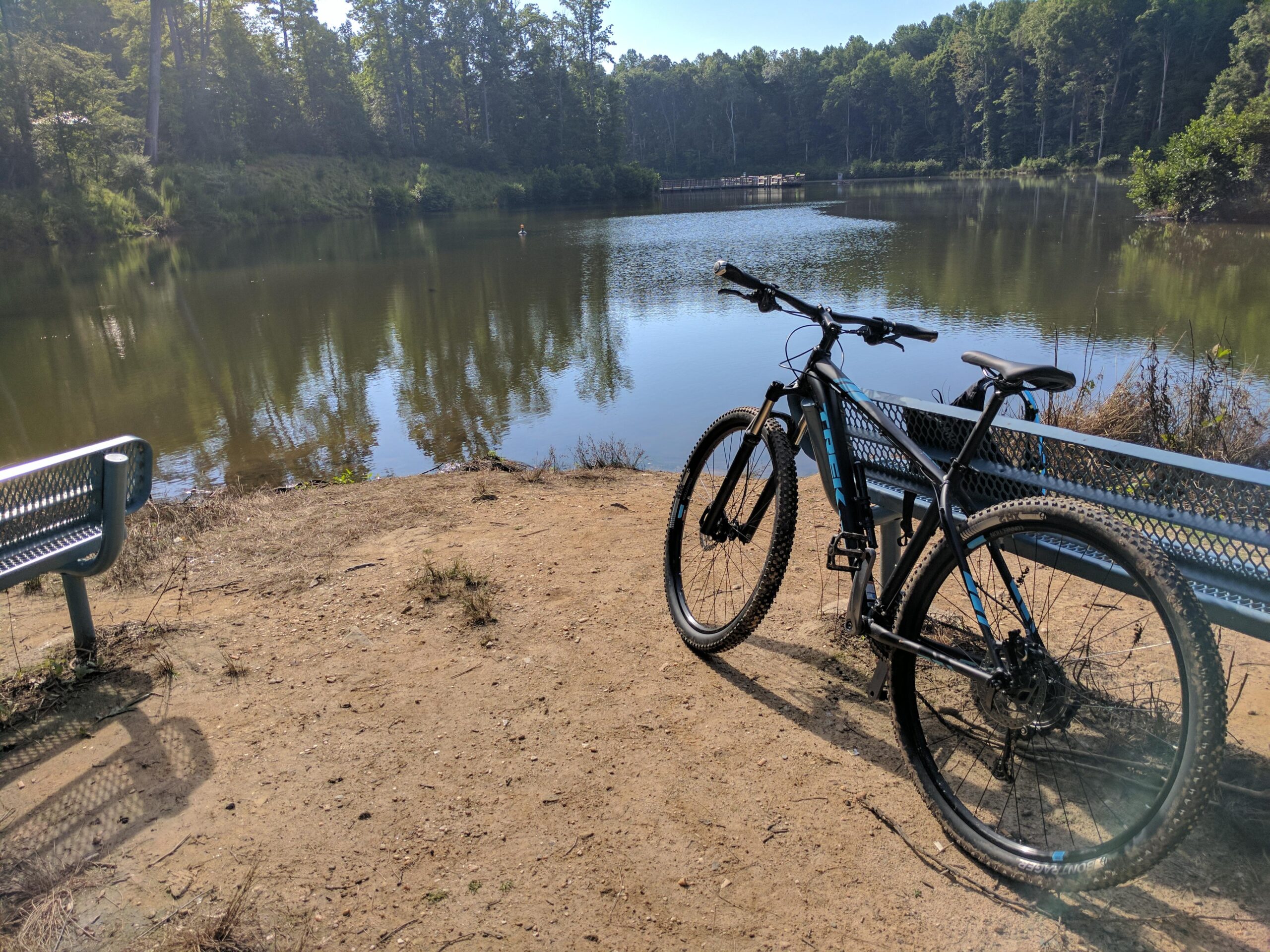 Trek X-Caliber 7: A serene outdoor scene featuring a mountain bike leaning against a metal bench beside a calm lake. The water reflects the greenery of surrounding trees, and a clear blue sky can be seen above. The area is peaceful, with a dirt path leading to the water's edge.