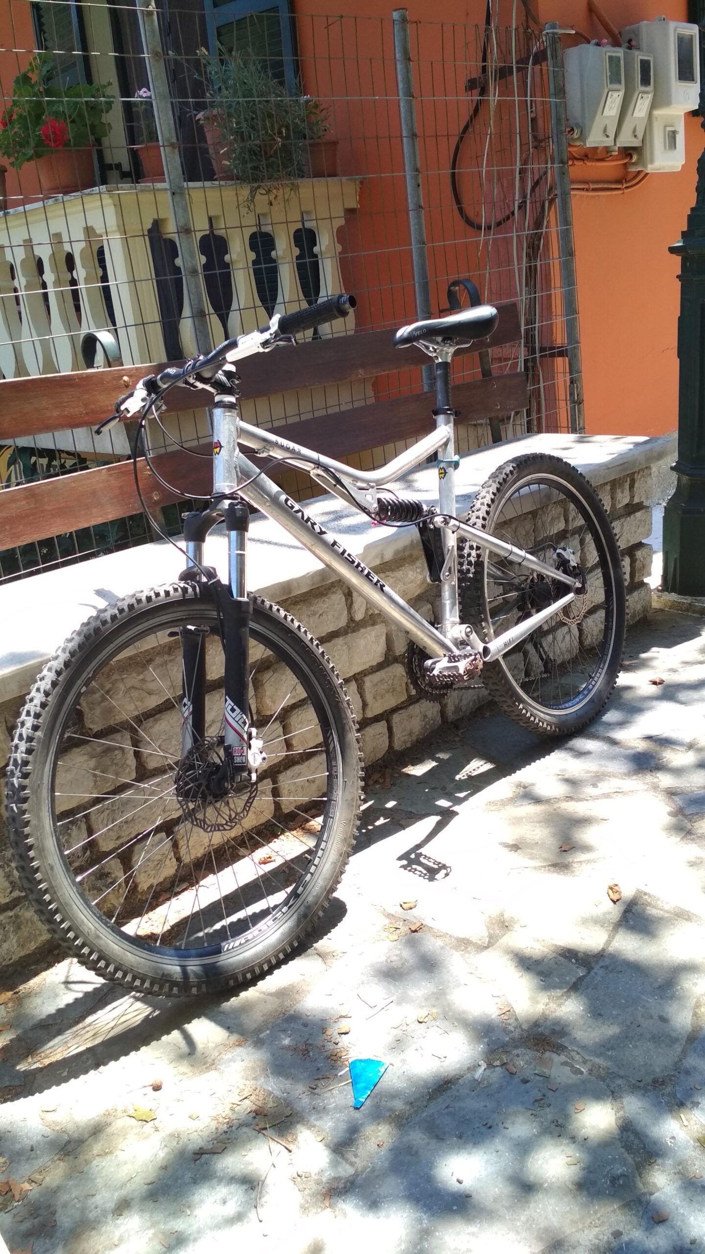 Gary Fisher HiFi: A mountain bike parked on a stone pathway next to a wooden bench, with a background featuring a fence and a building with potted plants. The bike has a silver frame and thick tires, and sunlight casts shadows on the ground.