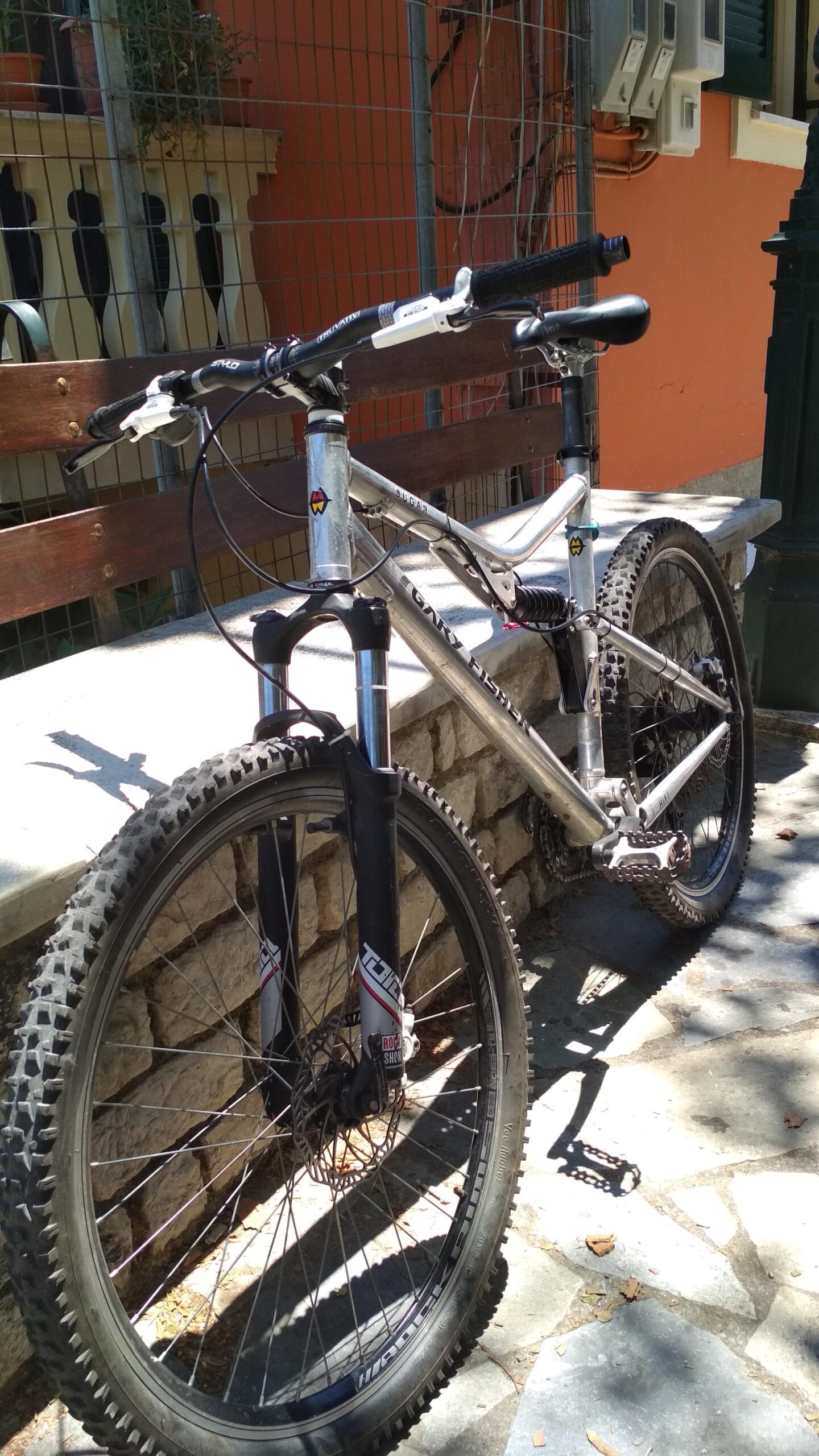 Gary Fisher HiFi: A close-up view of a mountain bike leaning against a stone wall. The bike features a silver aluminum frame, thick tires, and a front suspension fork. In the background, there is a blurred glimpse of a wooden bench and a colorful building, suggesting an outdoor setting. Sunlight casts shadows on the ground, creating a lively atmosphere.