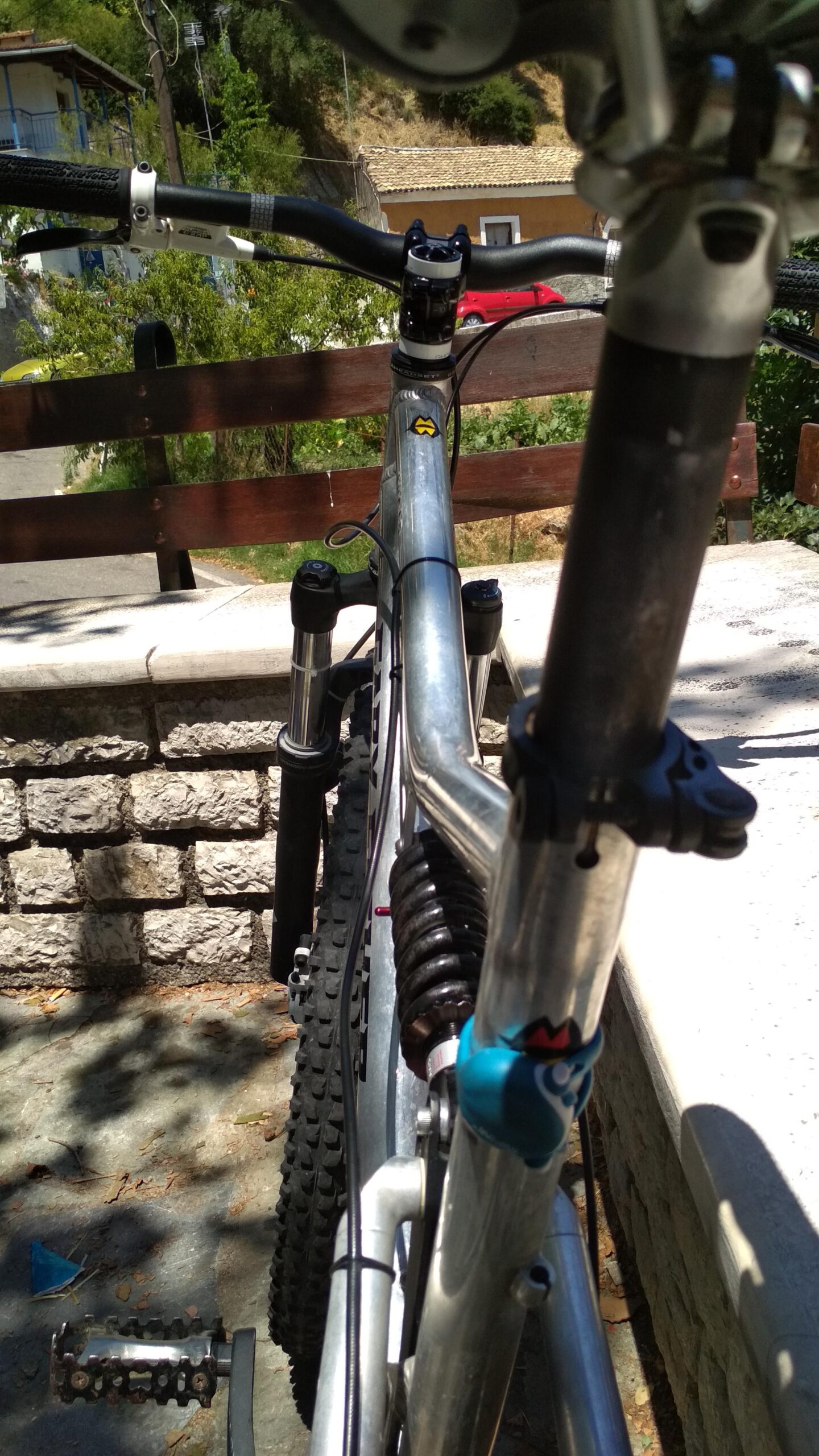 Gary Fisher HiFi: A close-up view of a mountain bike parked on a stone surface, showing the handlebars and front suspension. The bike has a metallic frame and a blue decorative element on the seat post. In the background, there are trees and a glimpse of a rural road with a house.