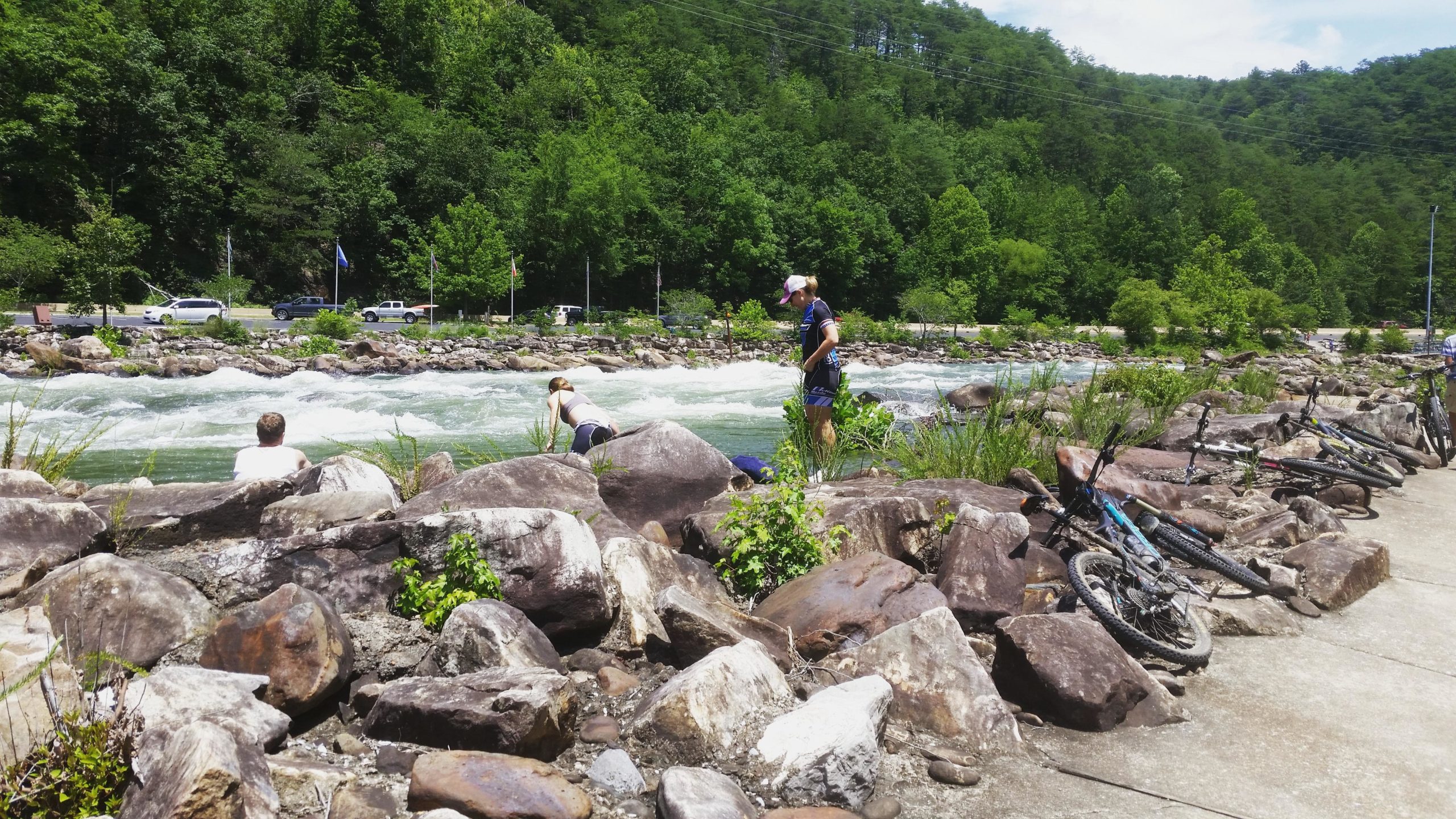 A scenic river view with people enjoying the outdoors. In the foreground, a person sits on large rocks near the water, while two individuals are standing by the riverbank. Several bicycles are leaned against the rocks, and lush green trees line the background. The water flows rapidly, indicating a recreational area suitable for activities like kayaking or fishing. Tanasi Trail System mountain bike trail.