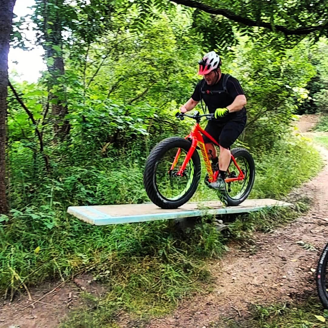 A person riding a bright red fat bike while performing a jump over a narrow wooden bridge in a lush green forested area. The cyclist is wearing a helmet and gloves and appears focused, with trees and a dirt path visible in the background. Olson Park mountain bike trail.