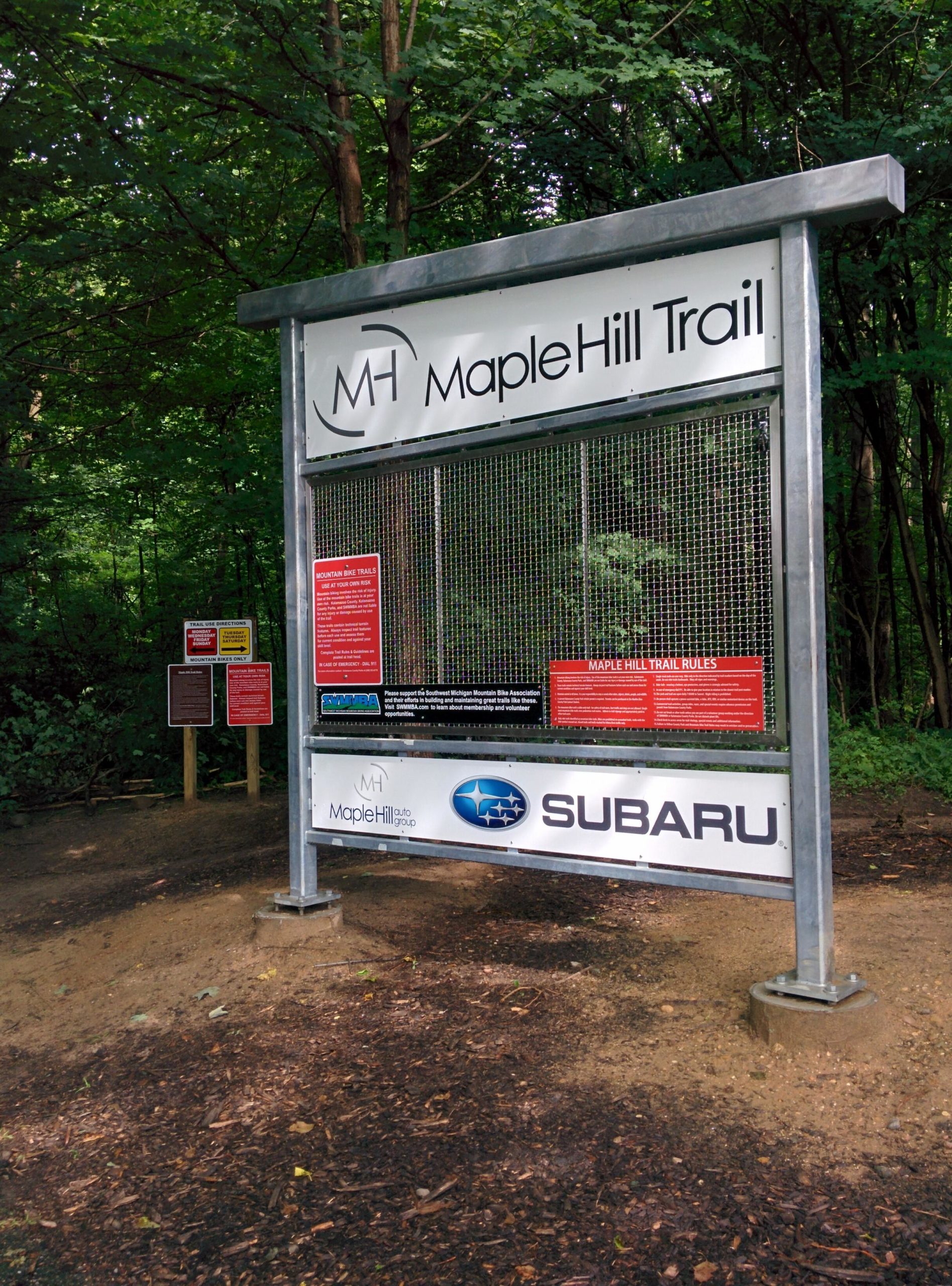 Sign marking the entrance to the Maple Hill Trail, featuring trail rules and informational notices. The sign is surrounded by dense trees, indicating a natural outdoor setting. Logos for Maple Hill and Subaru are displayed at the bottom of the sign. Maple Hill mountain bike trail.