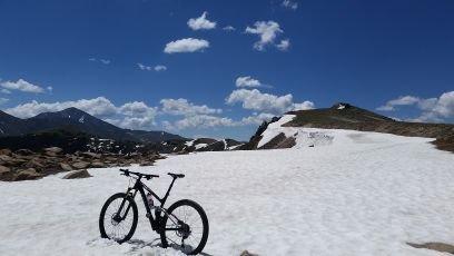 A mountain bike parked on a snow-covered landscape with mountains and a blue sky in the background, featuring scattered clouds. Monarch Crest Trail mountain bike trail.