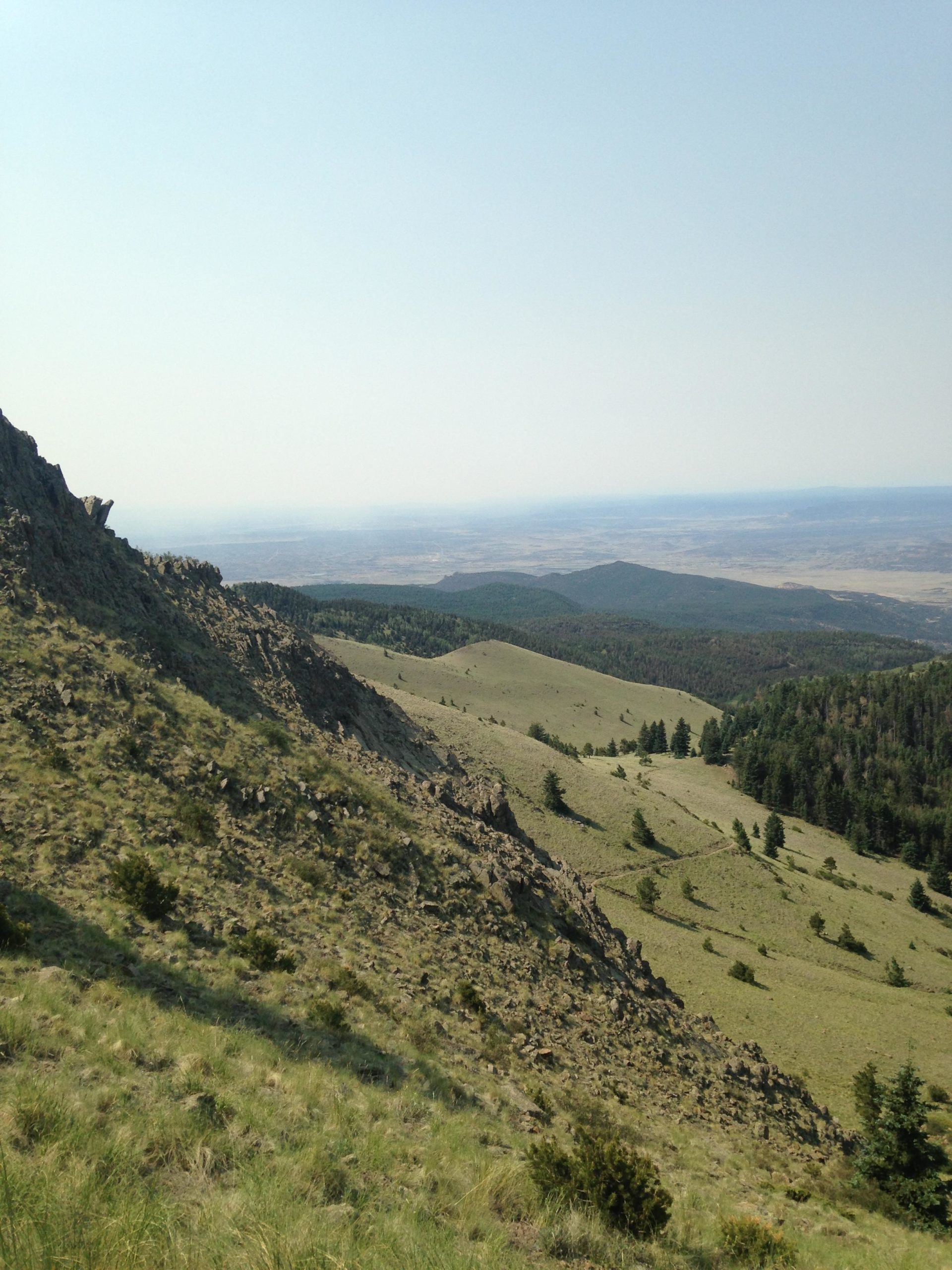 A panoramic view of a mountainous landscape featuring rolling hills, rocky outcrops, and patches of trees under a clear sky. The foreground shows grassy slopes leading down to a valley, while the horizon reveals distant mountains and flatlands. Mount Taylor Downhill mountain bike trail.