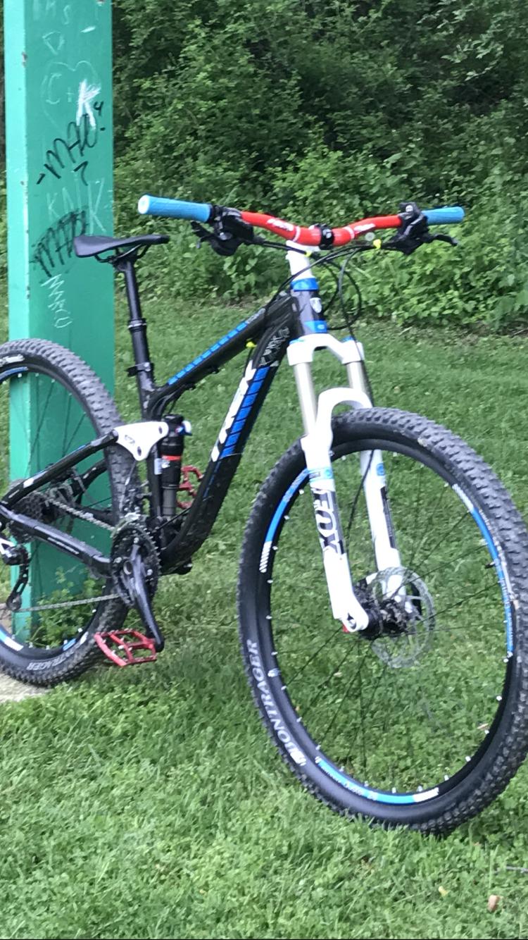 Trek Fuel EX 7: A black and blue mountain bike leaning against a green pole. The bike features thick tires suited for off-road terrain, with a white front fork and red and blue handlebars. In the background, there is a blur of green foliage and grass.