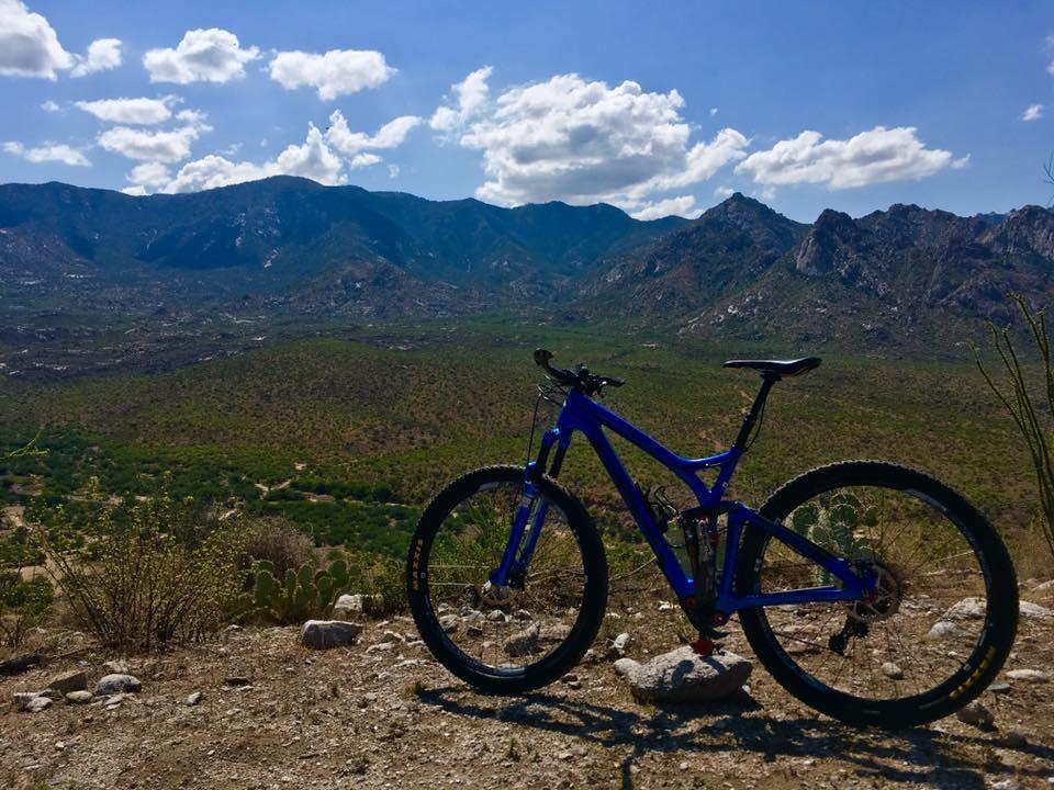 A vibrant blue mountain bike is parked on a rocky overlook, with a panoramic view of green valleys and rugged mountains in the background under a partly cloudy sky. 50-year Trail / Golder Ranch mountain bike trail.