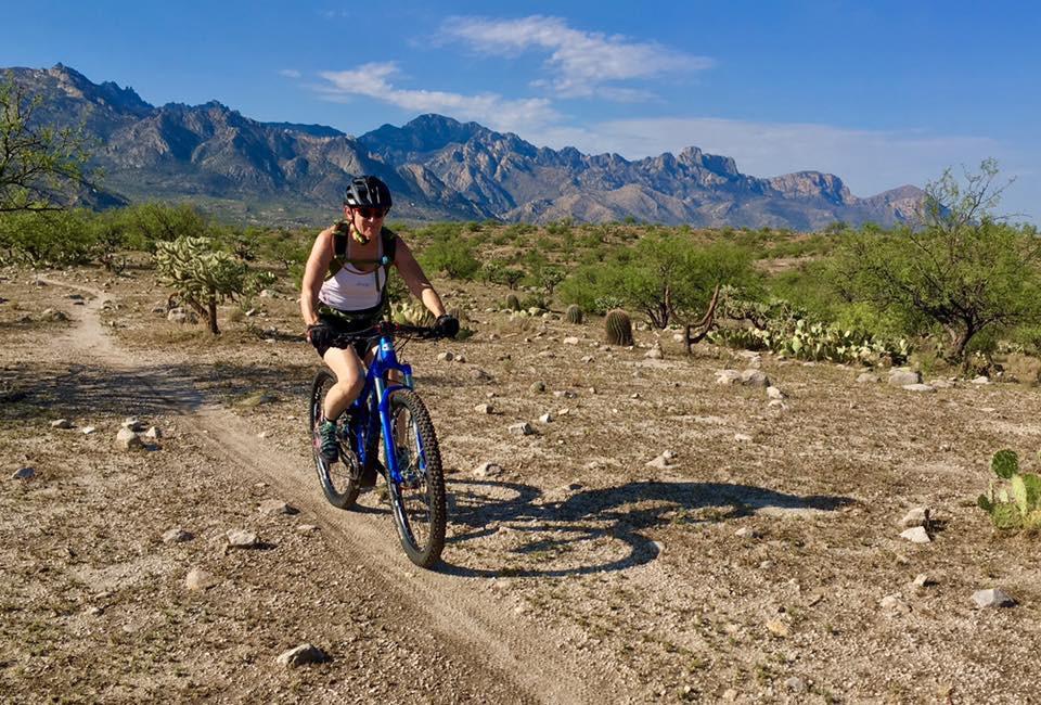A person riding a mountain bike along a dirt trail in a desert landscape, with rocky terrain and sparse vegetation. The background features distant mountains under a clear blue sky. 50-year Trail / Golder Ranch mountain bike trail.