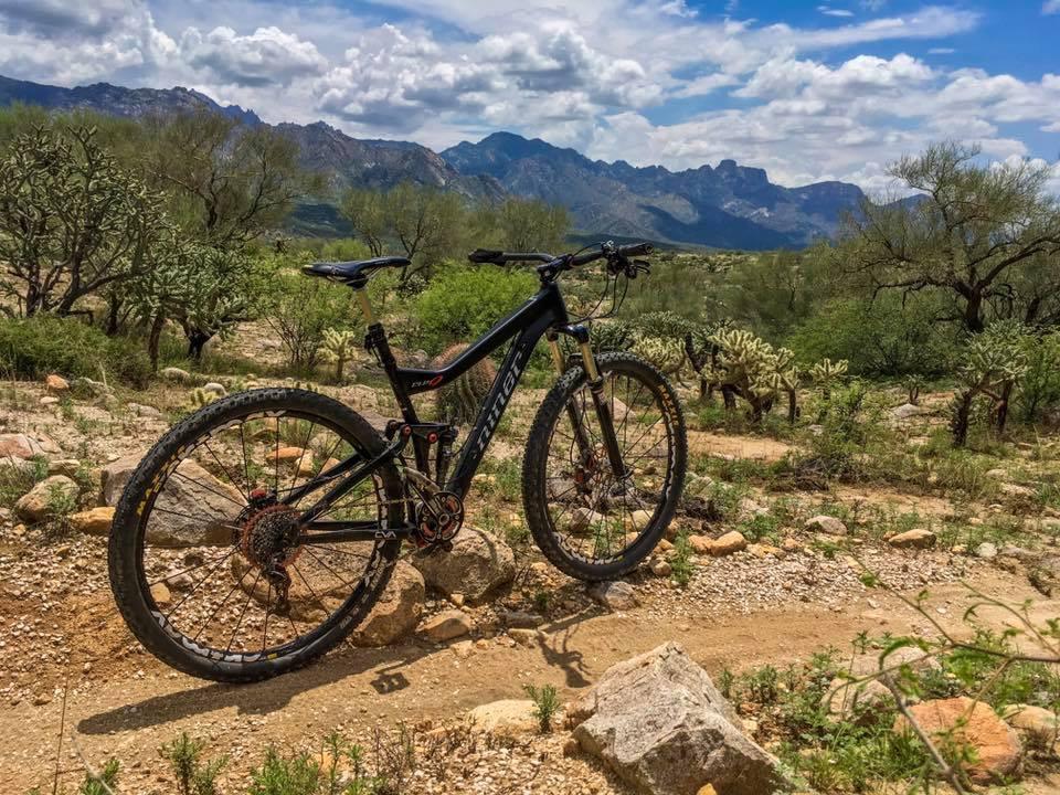 A mountain bike resting on a dirt trail surrounded by rocky terrain and desert vegetation, with a backdrop of mountains under a partly cloudy sky. 50-year Trail / Golder Ranch mountain bike trail.