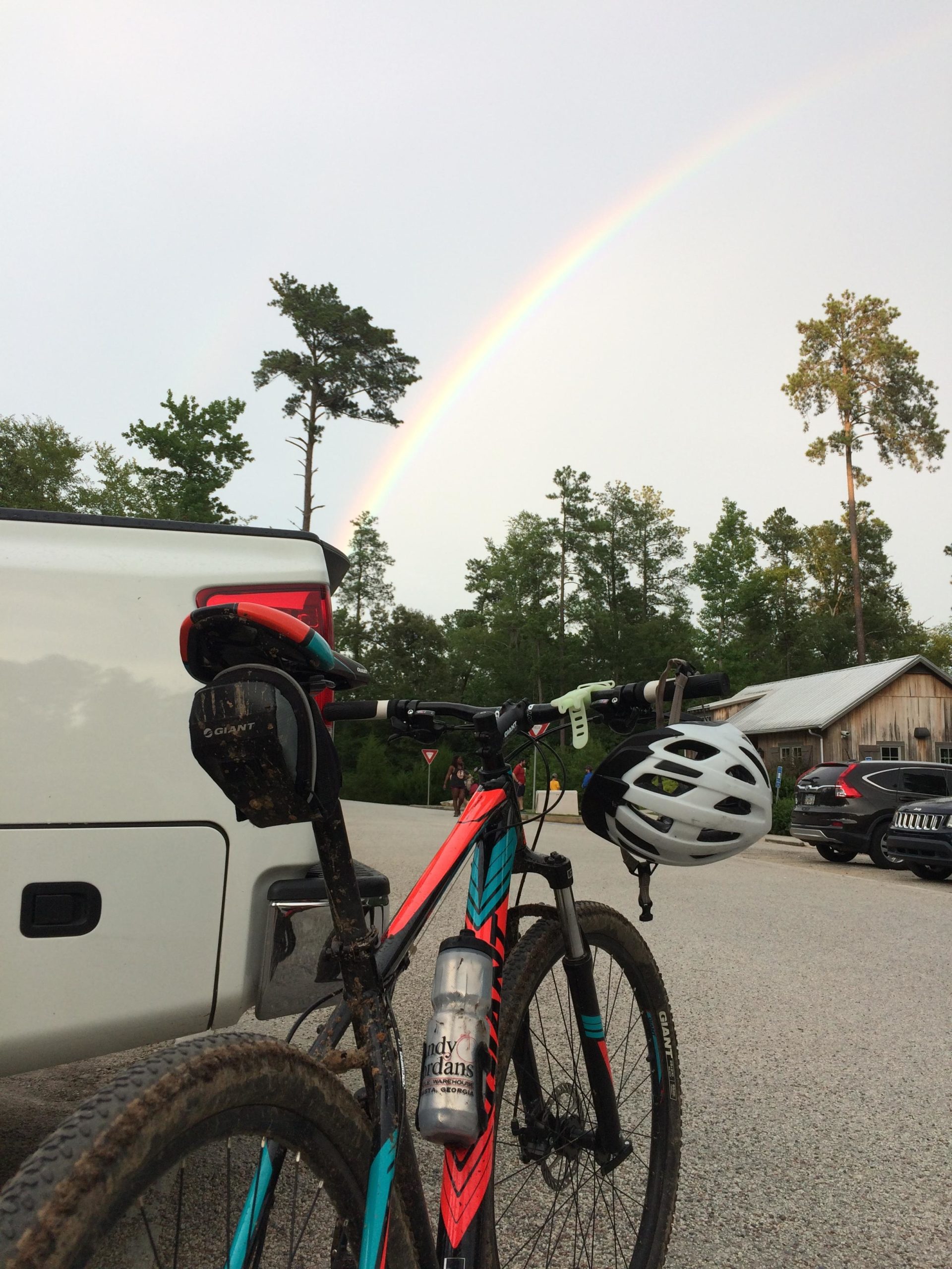 A close-up view of a brightly colored mountain bike with muddy tires, parked next to a white pickup truck. A rainbow arcs across the sky in the background, with green trees surrounding the area. A bicycle helmet hangs from the handlebars, and a water bottle is attached to the frame. Augusta Canal mountain bike trail.