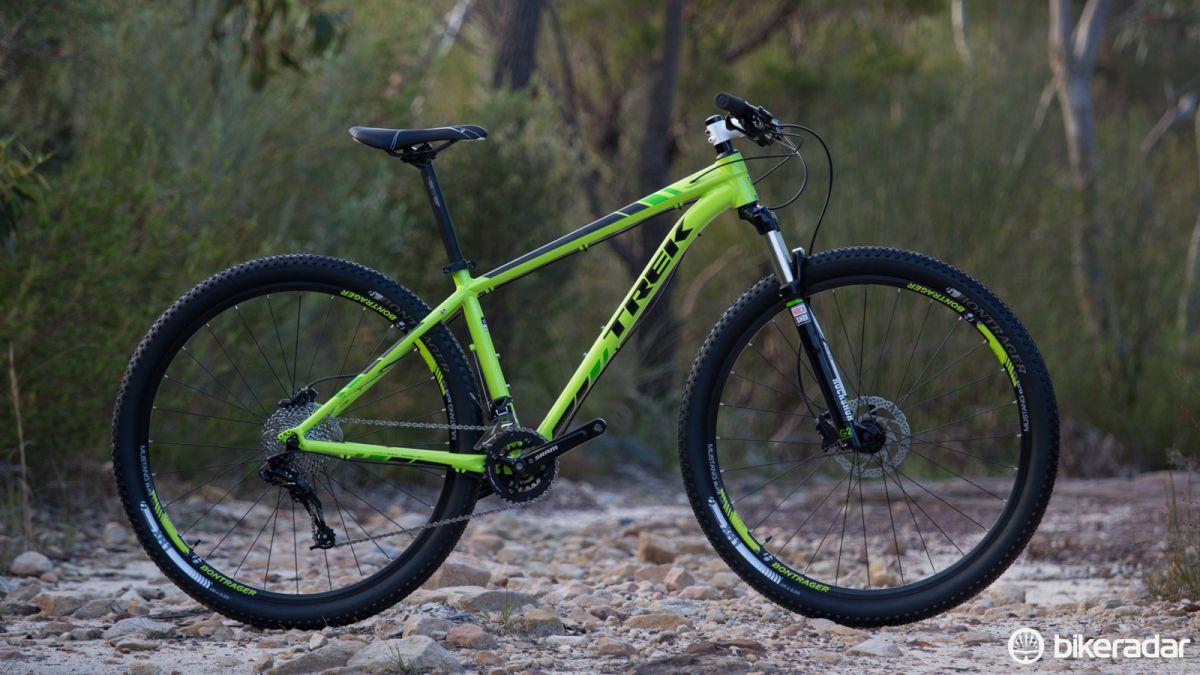 Trek Excalibur: A bright green mountain bike with black and white accents is positioned on rocky terrain, surrounded by greenery in the background. The bike features large, knobby tires and a lightweight frame, designed for off-road cycling.