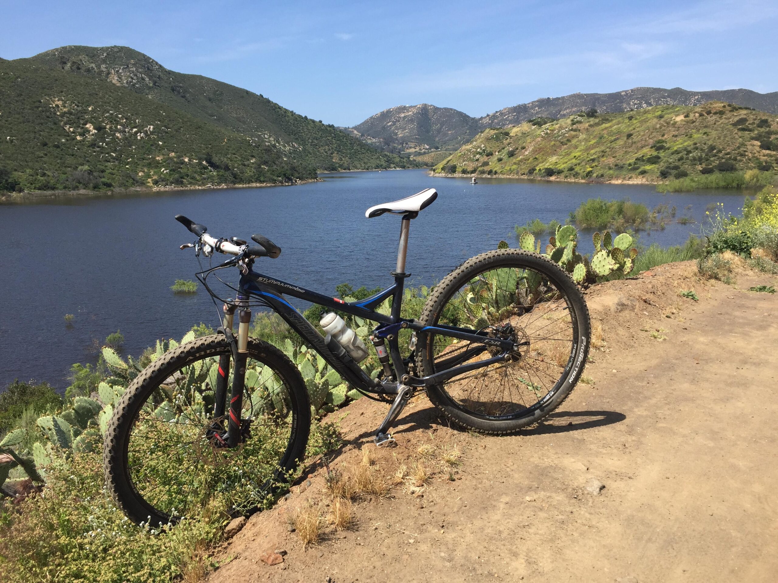 Specialized Stumpjumper FSR Comp 29er: A mountain bike parked on a dirt trail overlooking a serene lake surrounded by green hills and cacti under a clear blue sky.