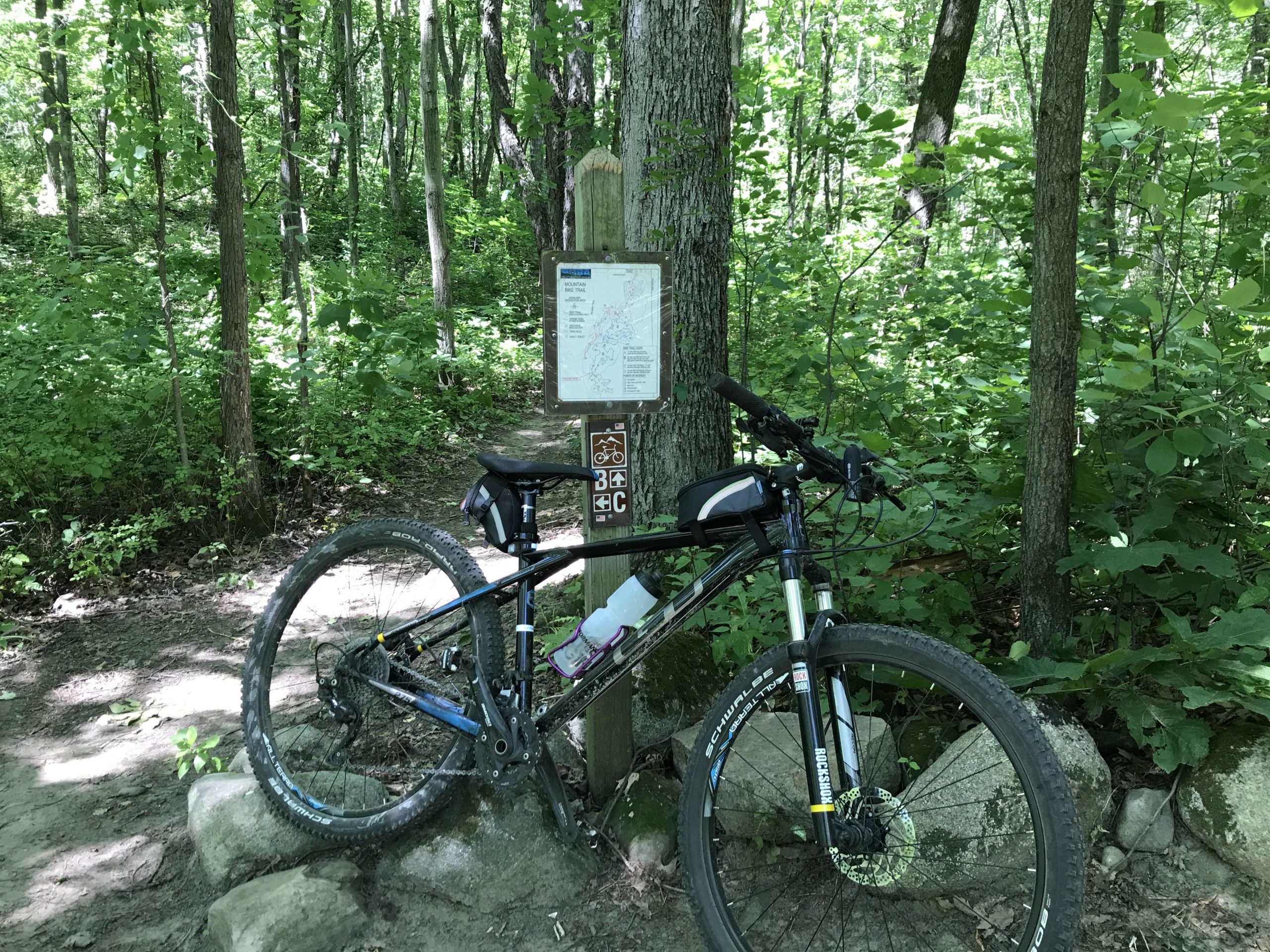 A mountain bike is parked against a tree near a marked trail sign in a lush green forest. The sign displays a trail map and information for cyclists. Surrounding the area are tall trees and dense foliage, with sunlight filtering through the leaves. Highland Recreation mountain bike trail.