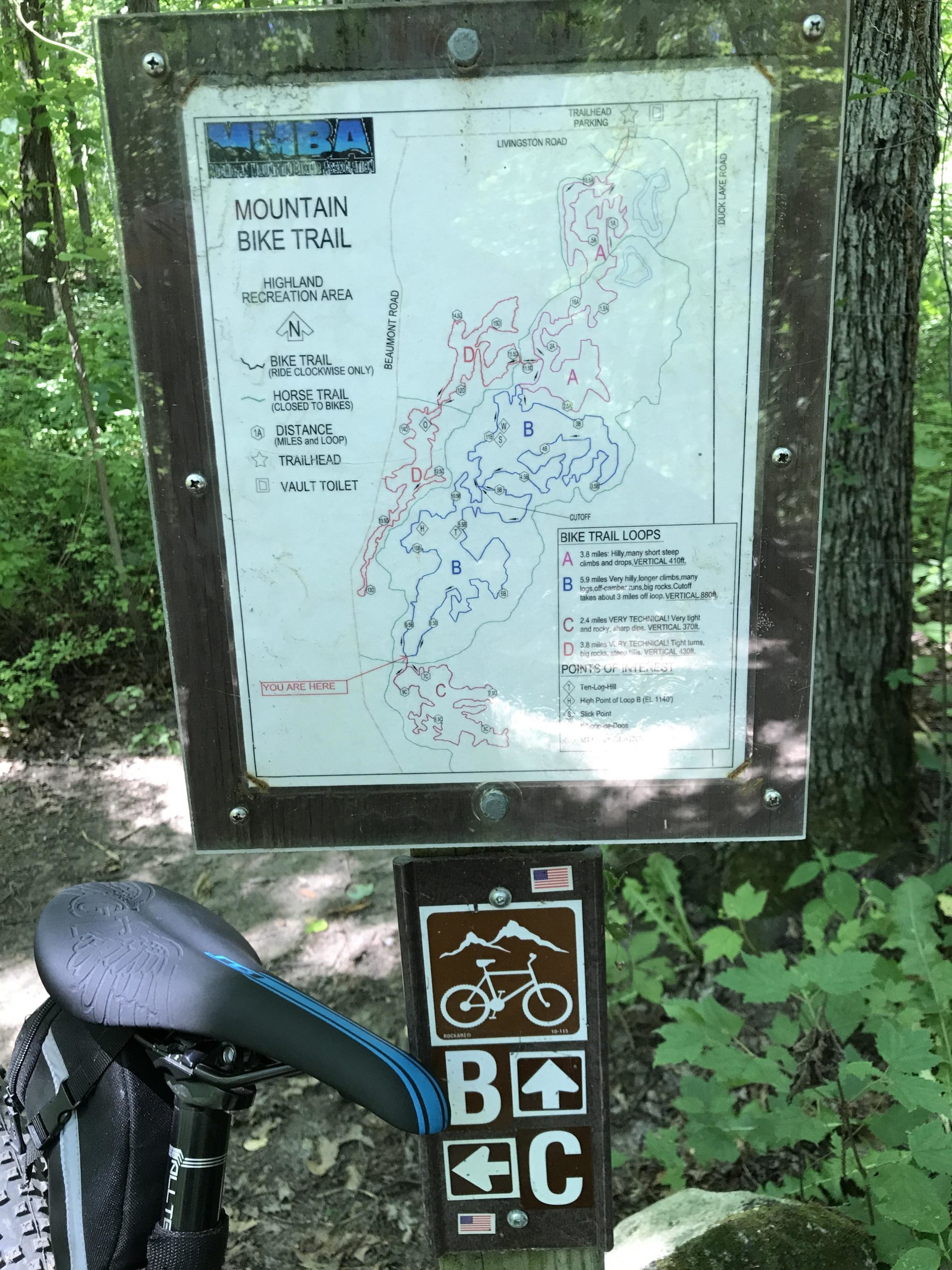 Sign for a mountain bike trail in a wooded area, featuring a map of the trails, including loops marked A, B, C, and D, with symbols indicating bike and horse trails. A bike seat and part of a backpack are visible in the foreground. The sign includes directional arrows and information about trailhead parking and distances. Highland Recreation mountain bike trail.