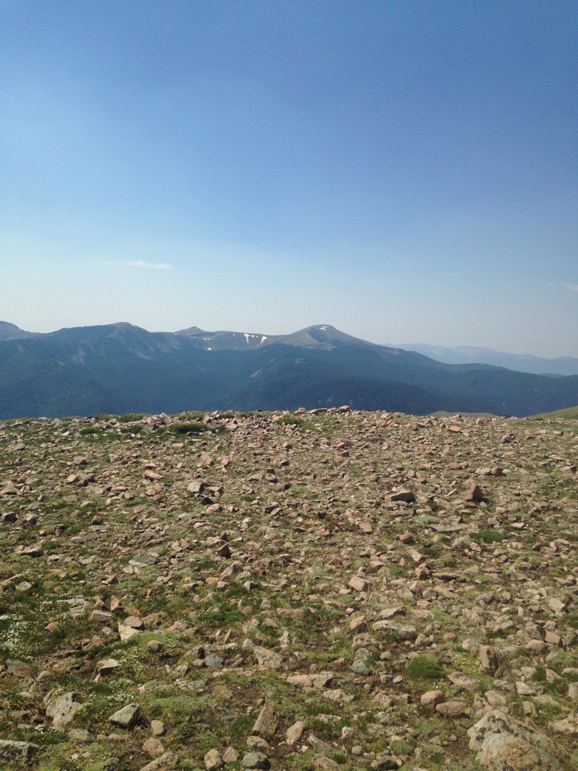 A scenic view of a mountainous landscape featuring rolling hills and peaks under a clear blue sky. The foreground is covered with rocky terrain and patches of grass, while the distant mountains show hints of snow on their summits. Northside At Taos Ski Valley mountain bike trail.