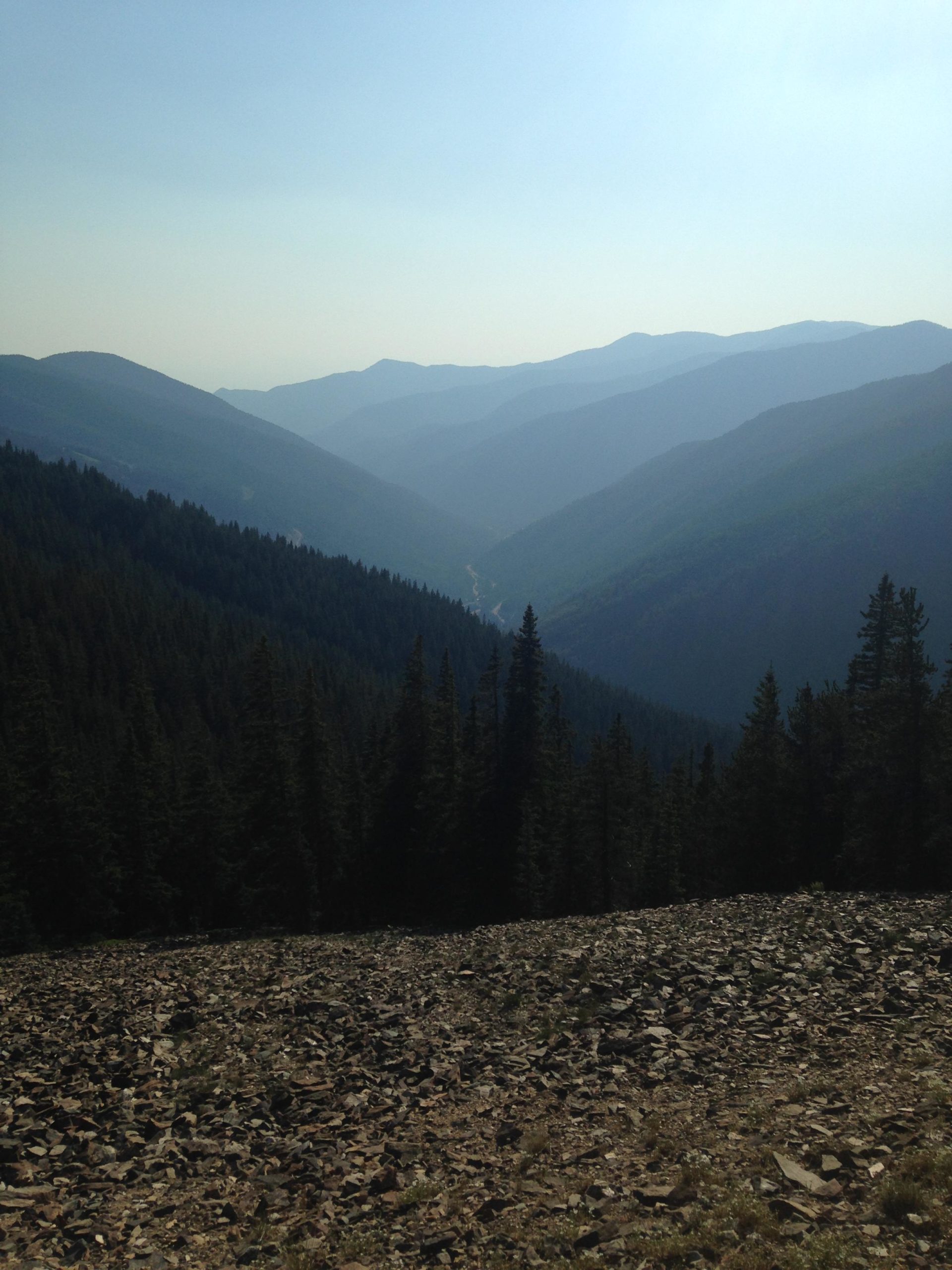 A panoramic view of a mountain valley, showcasing rolling hills in the distance, a winding river visible between the peaks, and a foreground of rocky terrain with scattered stones. The scene is bathed in soft, hazy light, giving it a tranquil and serene atmosphere. Dense evergreen forests line the lower slopes of the mountains. Northside At Taos Ski Valley mountain bike trail.