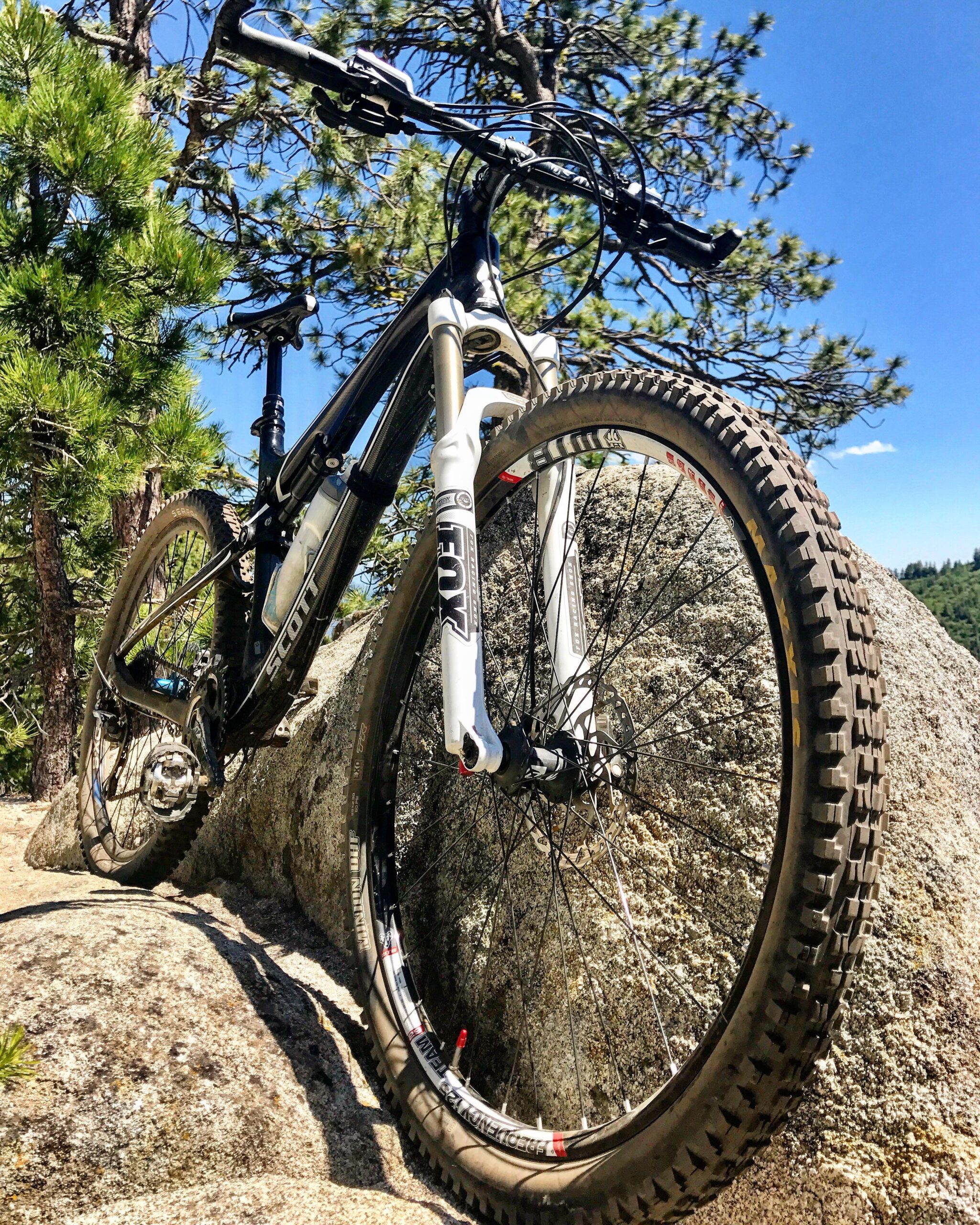 Scott Genius 920: A mountain bike positioned on a large rock, surrounded by trees and a clear blue sky. The bike is angled to showcase its tires and frame, highlighting its features and rugged design.