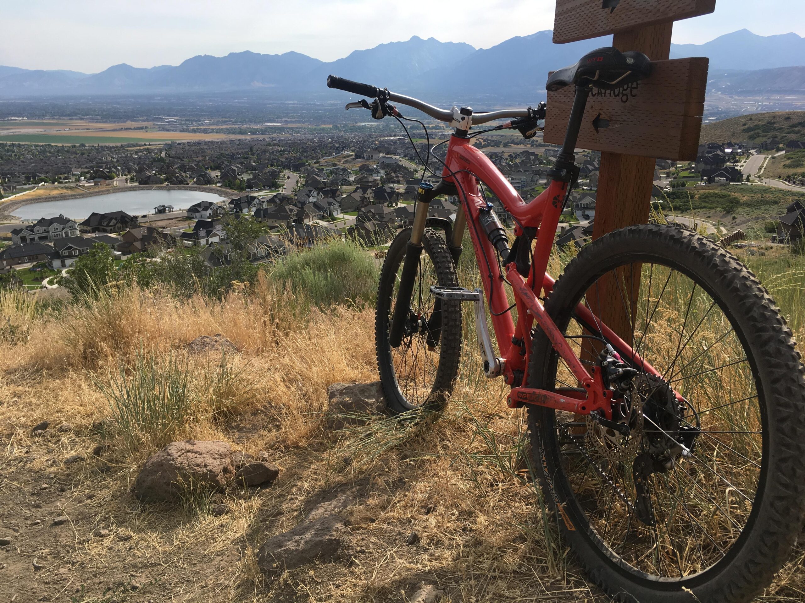 Santa Cruz Nomad: A bright red mountain bike leaning against a wooden trail sign, overlooking a scenic valley filled with houses and features. In the background, rolling hills and mountains are visible under a clear sky. The foreground includes dry grass and some rocky terrain.